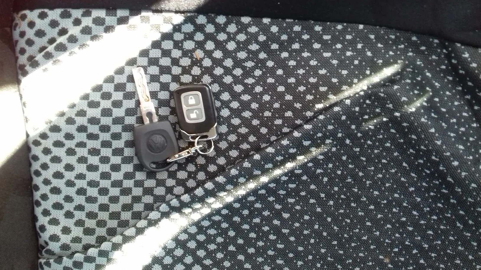 Kluczyk do samochodu marki Volkswagen z pilotem do zdalnego sterowania, leżący na szarej, wzorzystej tapicerce samochodowej w kratkę. Pilot posiada dwa przyciski: jeden z ikoną otwartej kłódki i drugi z ikoną zamkniętej kłódki.
