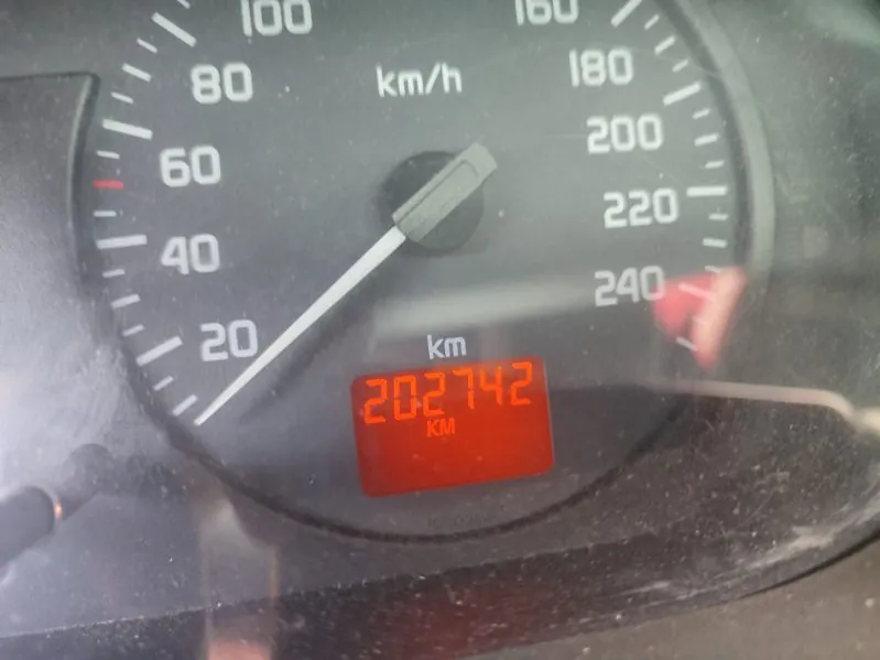 Zbliżenie na zegar samochodowy. Wskazówka prędkościomierza wskazuje około 20 km/h. Widoczna skala prędkości od 0 do 240 km/h z oznaczeniem 'km/h' powyżej wskazówki. Poniżej znajduje się czerwony wyświetlacz cyfrowy z liczbą '202742' oraz napisem 'km' i 'KM' pod liczbą. Tło zegara jest ciemne, a szkło pokryte jest drobnymi zabrudzeniami.