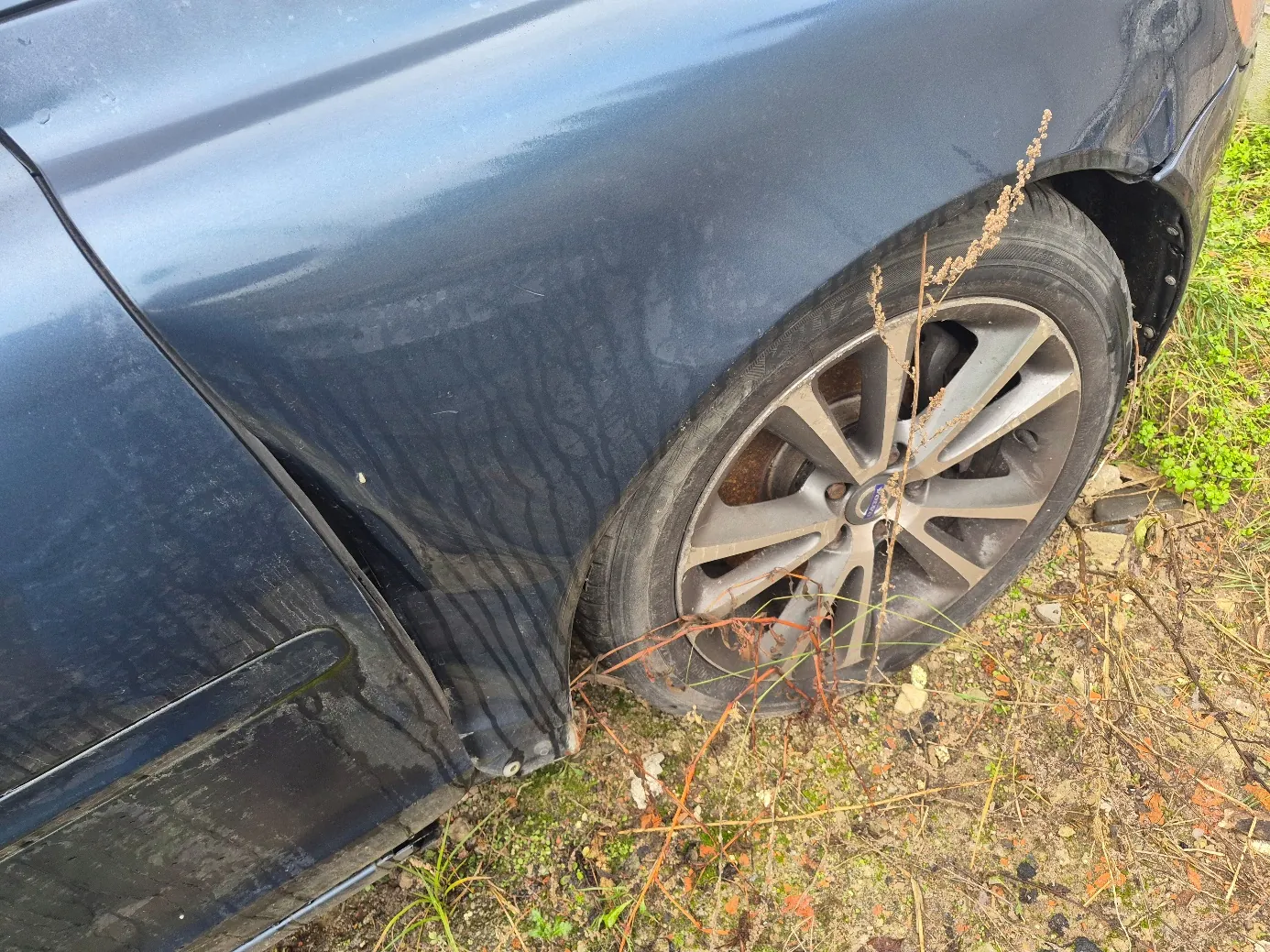 Czarny samochód widziany z boku, z bliska przedniego prawego koła. Koło ma srebrną felgę wieloramienną, a opona jest brudna. Na karoserii widoczne są zadrapania i uszkodzenia w okolicy nadkola. Pod samochodem i wokół niego rośnie trawa oraz sucha roślinność na brudnej, ziemistej powierzchni. W tle widoczne są zielone rośliny.