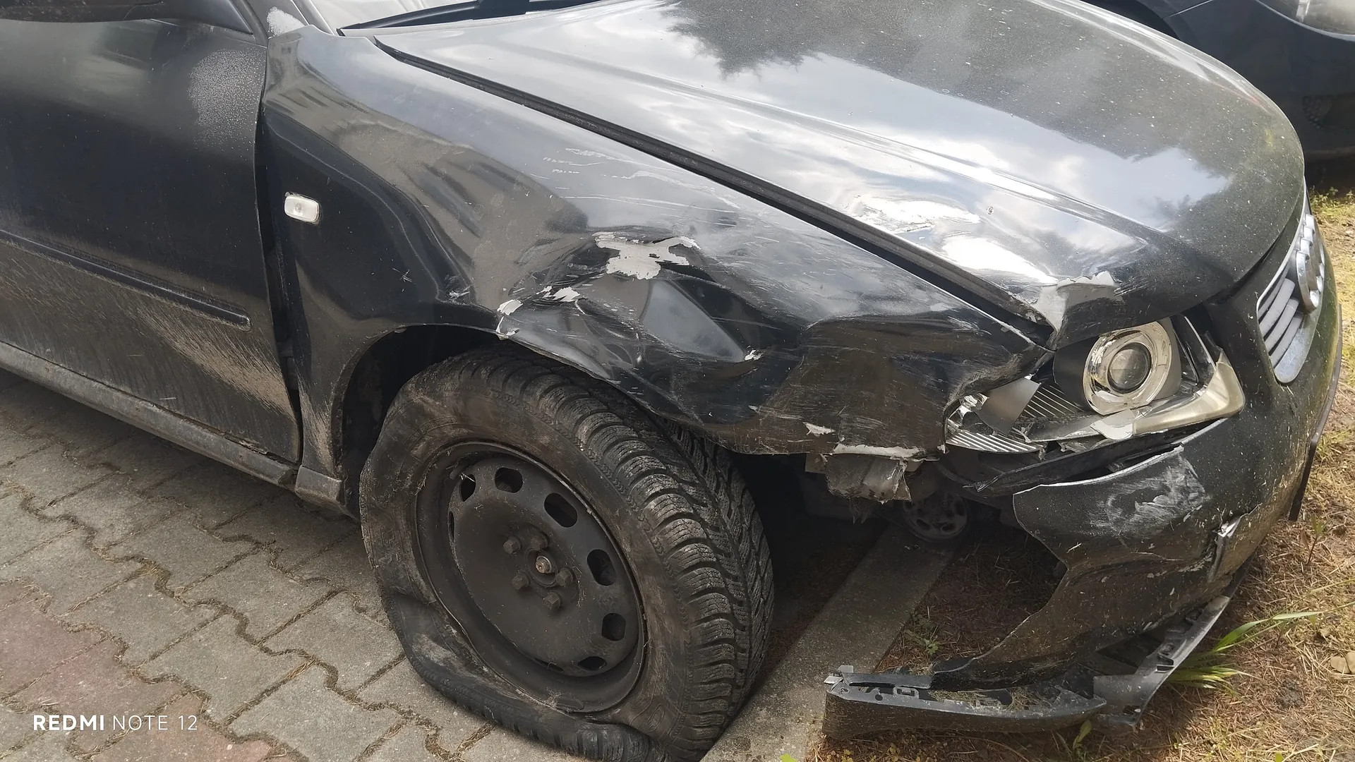 Zniszczony czarny samochód osobowy z widocznymi uszkodzeniami na przedniej lewej stronie: pomarszczona maska, pęknięte reflektory, uszkodzony zderzak oraz spłaszczona opona. Samochód stoi na brukowanej powierzchni, obok trawy. W lewym dolnym rogu widoczny napis 'REDMI NOTE 12'.