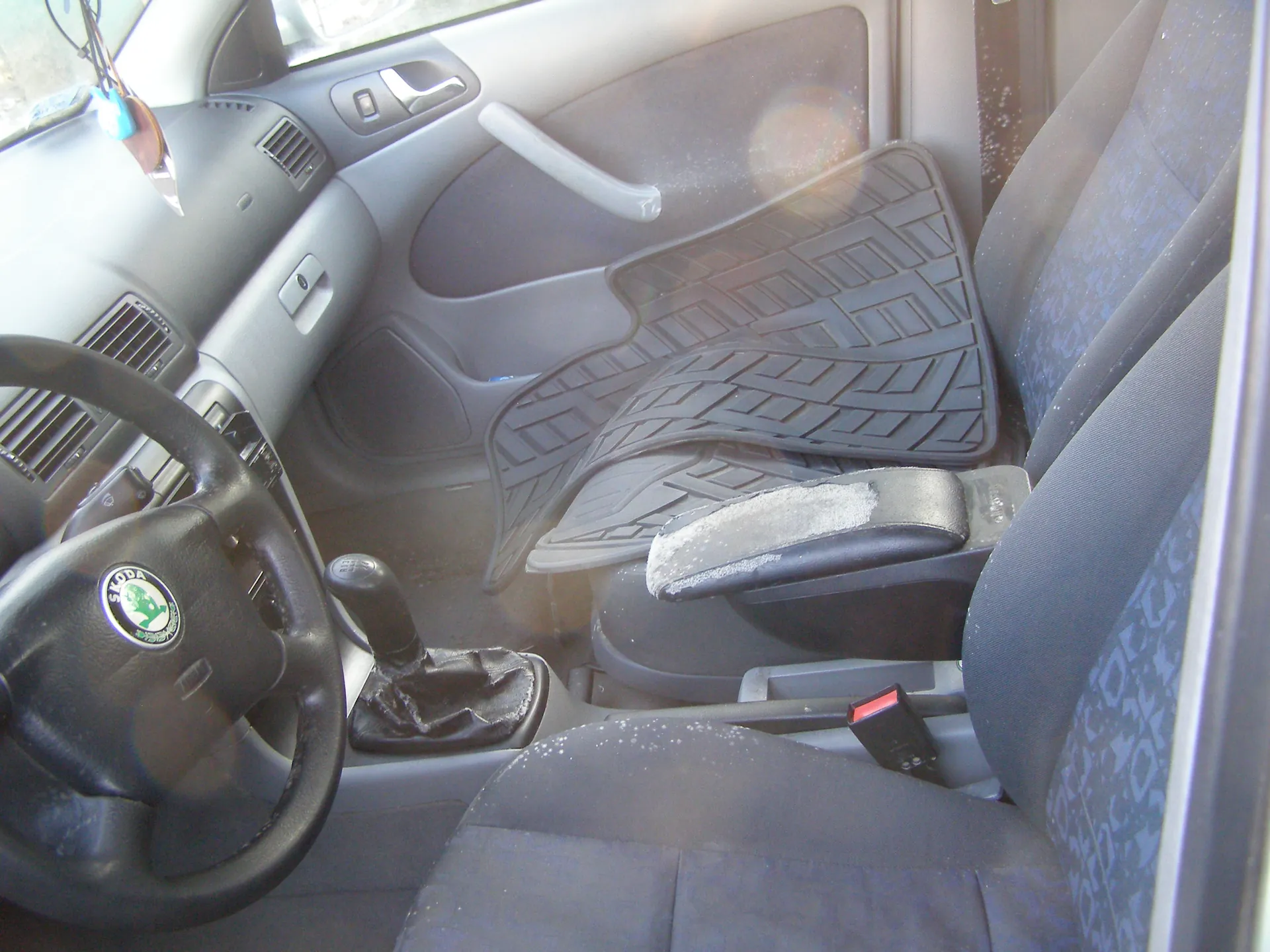 Wnętrze samochodu marki Škoda. Widoczna kierownica z logo Škoda, deska rozdzielcza z wentylacjami, dźwignia zmiany biegów, czarne gumowe dywaniki podłogowe z geometrycznym wzorem, siedzenia tapicerowane szarym materiałem z niebieskim wzorem. Na siedzeniach i podłodze widoczny jest śnieg. Drzwi pasażera z uchwytem i oknem. Wnętrze oświetlone światłem dziennym.