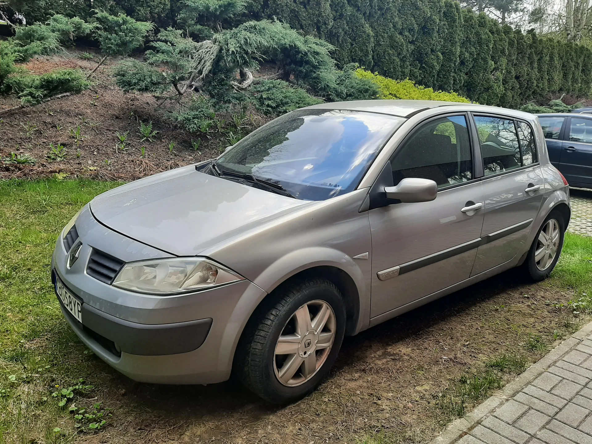 Srebrny samochód osobowy marki Renault zaparkowany na trawie. Widoczna tablica rejestracyjna HJ 58YP. W tle zielone krzewy, drzewa oraz fragment innego pojazdu. Samochód znajduje się na obszarze z trawą i częściowo odsłoniętą ziemią, obok chodnika z kostki.