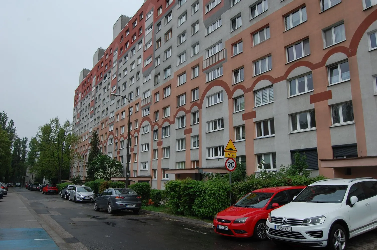 Wielopiętrowy blok mieszkalny o elewacji w kolorach szarym i pomarańczowym z licznymi oknami. Przed budynkiem znajduje się ulica z zaparkowanymi samochodami: białym SUV-em, czerwonym samochodem osobowym oraz szarym samochodem. Po prawej stronie widoczny jest znak drogowy ograniczający prędkość do 20 km/h z tabliczką '20 m'. Wzdłuż budynku rosną zielone krzewy i drzewa.