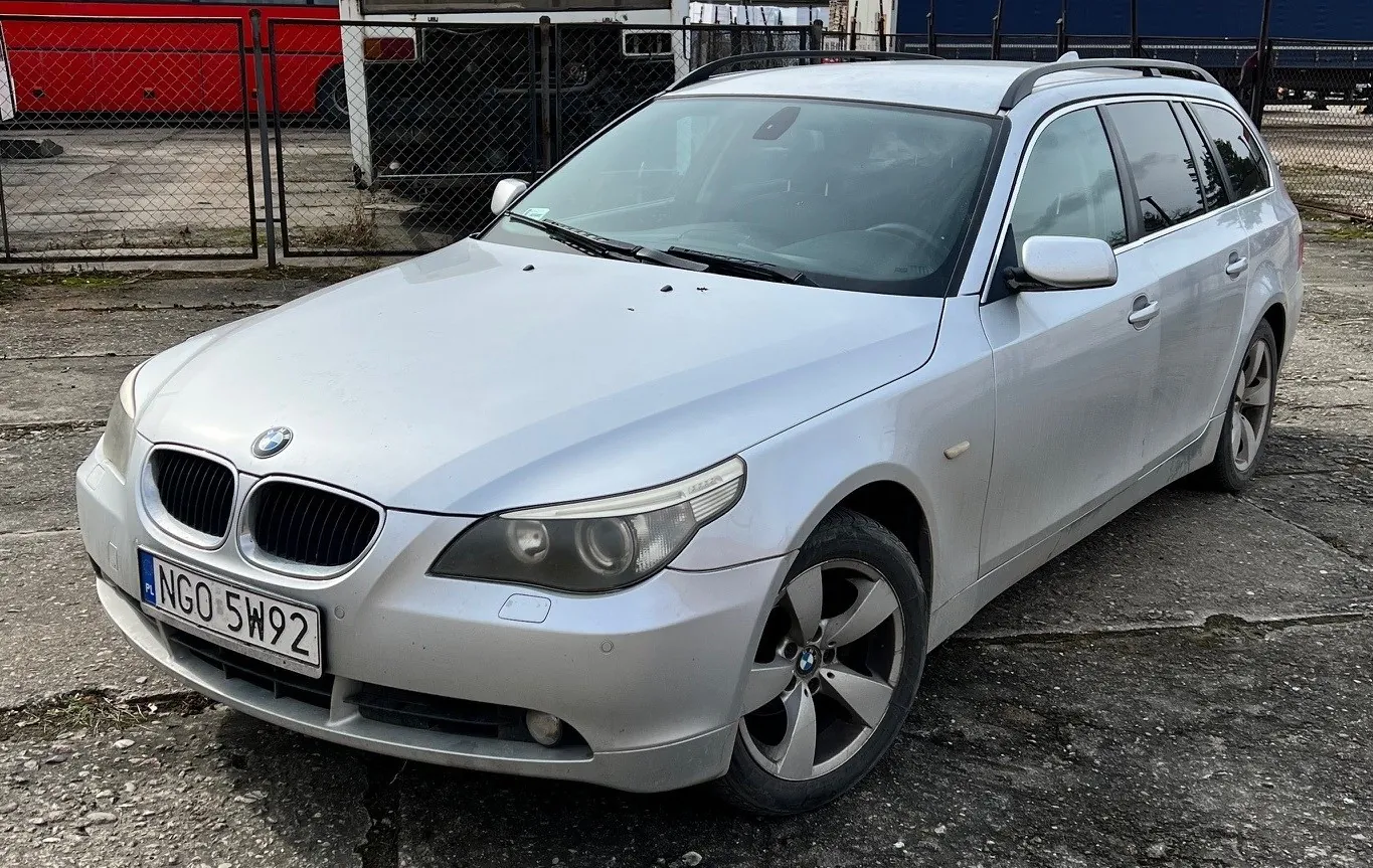 Srebrny samochód osobowy typu kombi marki BMW z tablicą rejestracyjną PL NGO 5W92. Pojazd stoi na betonowej powierzchni z widocznymi pęknięciami. W tle czarny płot siatkowy, czerwony pojazd oraz przemysłowe budynki. Na masce widoczny logo BMW, a na przednim zderzaku tablica rejestracyjna z białym tłem i czarnym napisem.