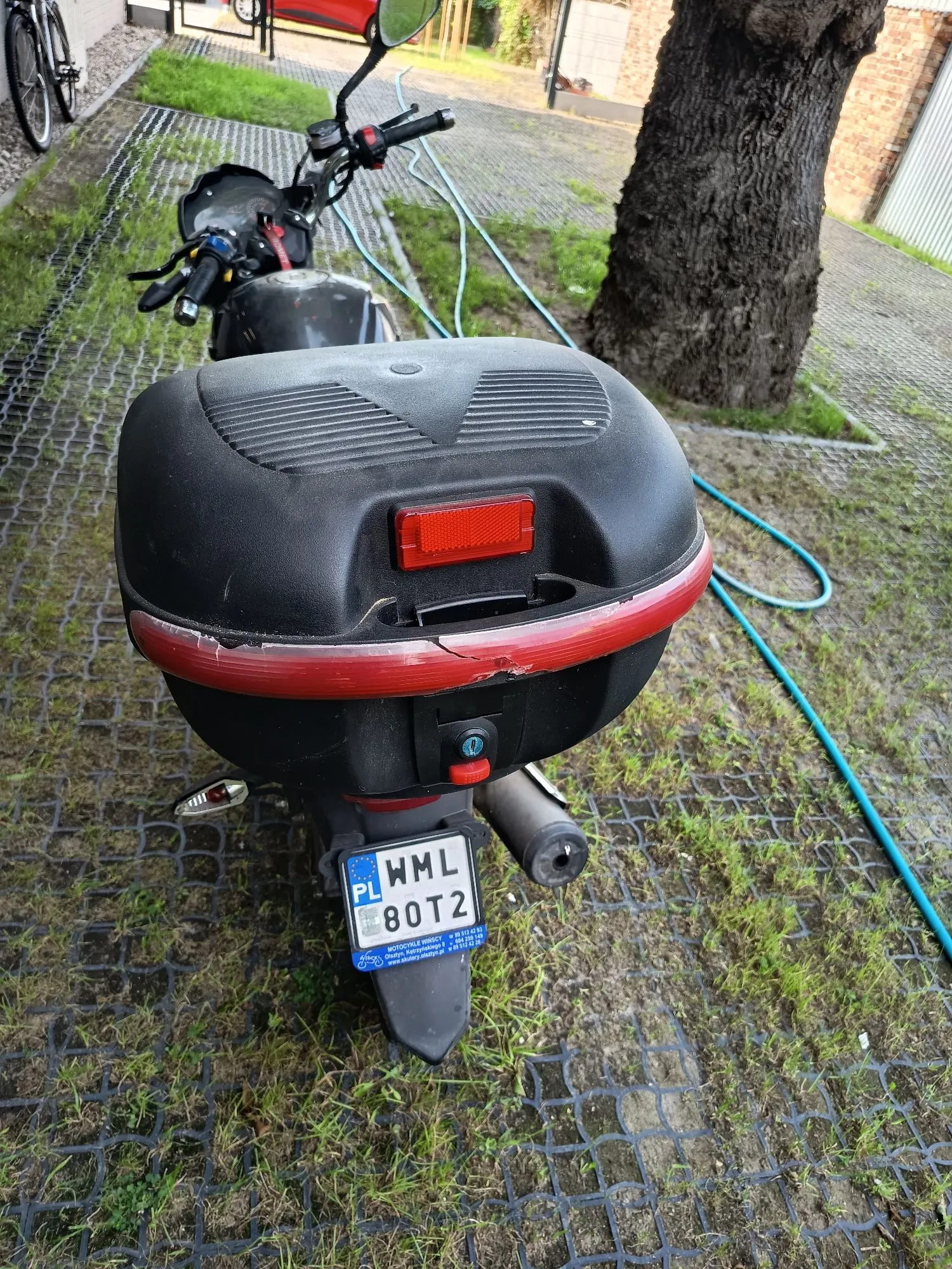 Widok z tyłu na czarny motocykl z zamontowanym dużym, czarnym kufrem. Kufer posiada czerwony odblask na górze oraz pęknięcie wzdłuż czerwonego paska w dolnej części. Poniżej znajduje się polska tablica rejestracyjna o numerze WML 80T2. Motocykl stoi na podłożu z plastikowych kratownic trawnikowych, obok znajduje się pień drzewa i niebieski wąż ogrodowy.