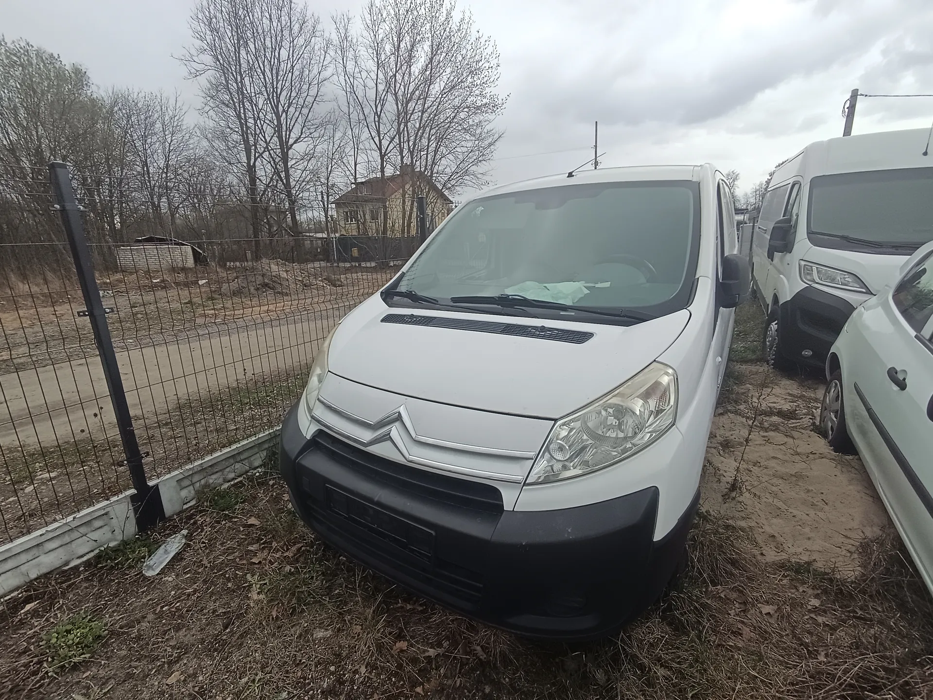 Biały samochód dostawczy Citroën na pierwszym planie, zaparkowany na suchym trawniku. Po prawej stronie widoczne są kolejne pojazdy: biały dostawczy i częściowo widoczny biały osobowy. Po lewej stronie metalowe ogrodzenie, za którym droga i suche drzewa. W tle dom jednorodzinny, bezlistne drzewa oraz pochmurne niebo. Brak czytelnego tekstu na obrazie.