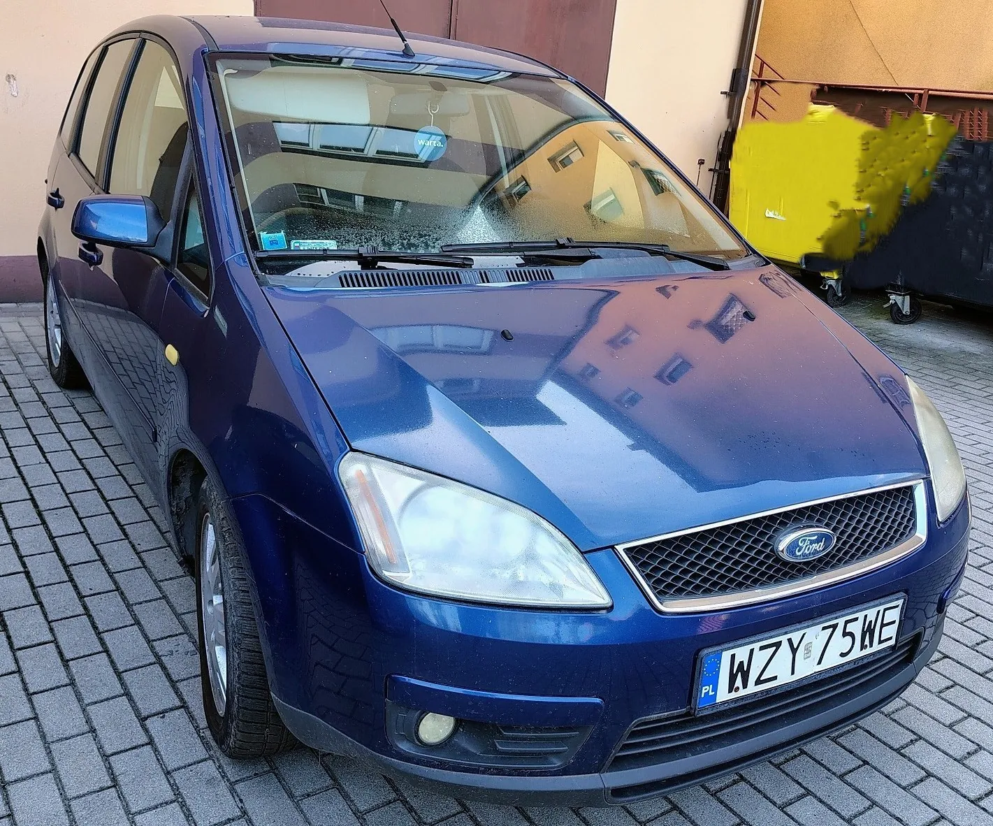 Niebieski samochód marki Ford zaparkowany na kostce brukowej. Widoczny jest przód pojazdu z tablicą rejestracyjną o numerze WZY 75WE. Samochód stoi przed jasną ścianą budynku.