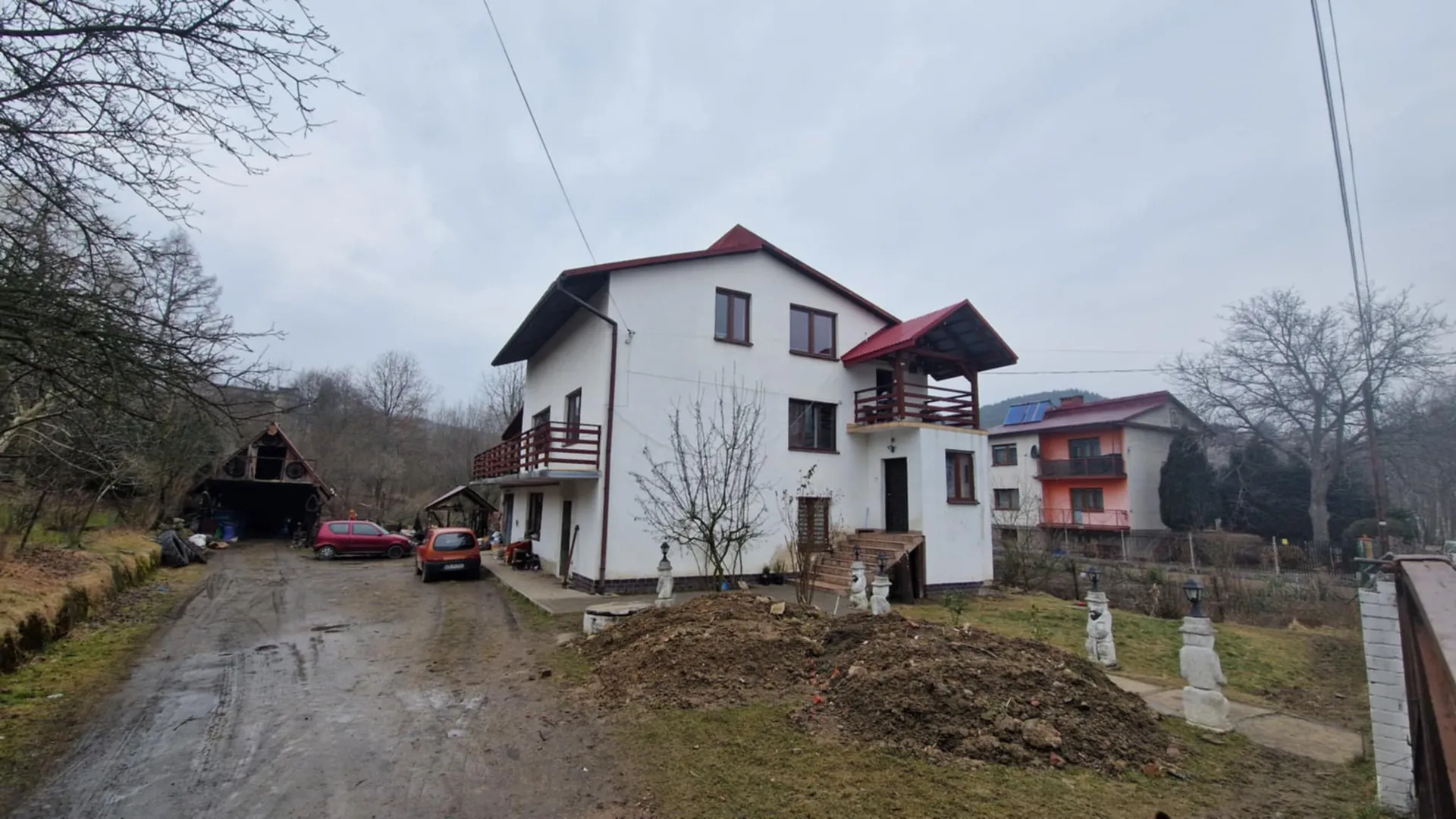 Biały dom dwupiętrowy z czerwonym dachem i drewnianymi balkonami, położony przy brudnej drodze ziemnej. Przy domu zaparkowane są dwa samochody: czerwony i pomarańczowy. Po lewej stronie widoczna drewniana budowla z otwartym wejściem, a w tle dom pomarańczowo-biały z czerwonym dachem i panelami słonecznymi. Na pierwszym planie stosy ziemi, nagie drzewa oraz kamienne filary z latarniami. Tło stanowią wzgórza i pochmurne niebo.