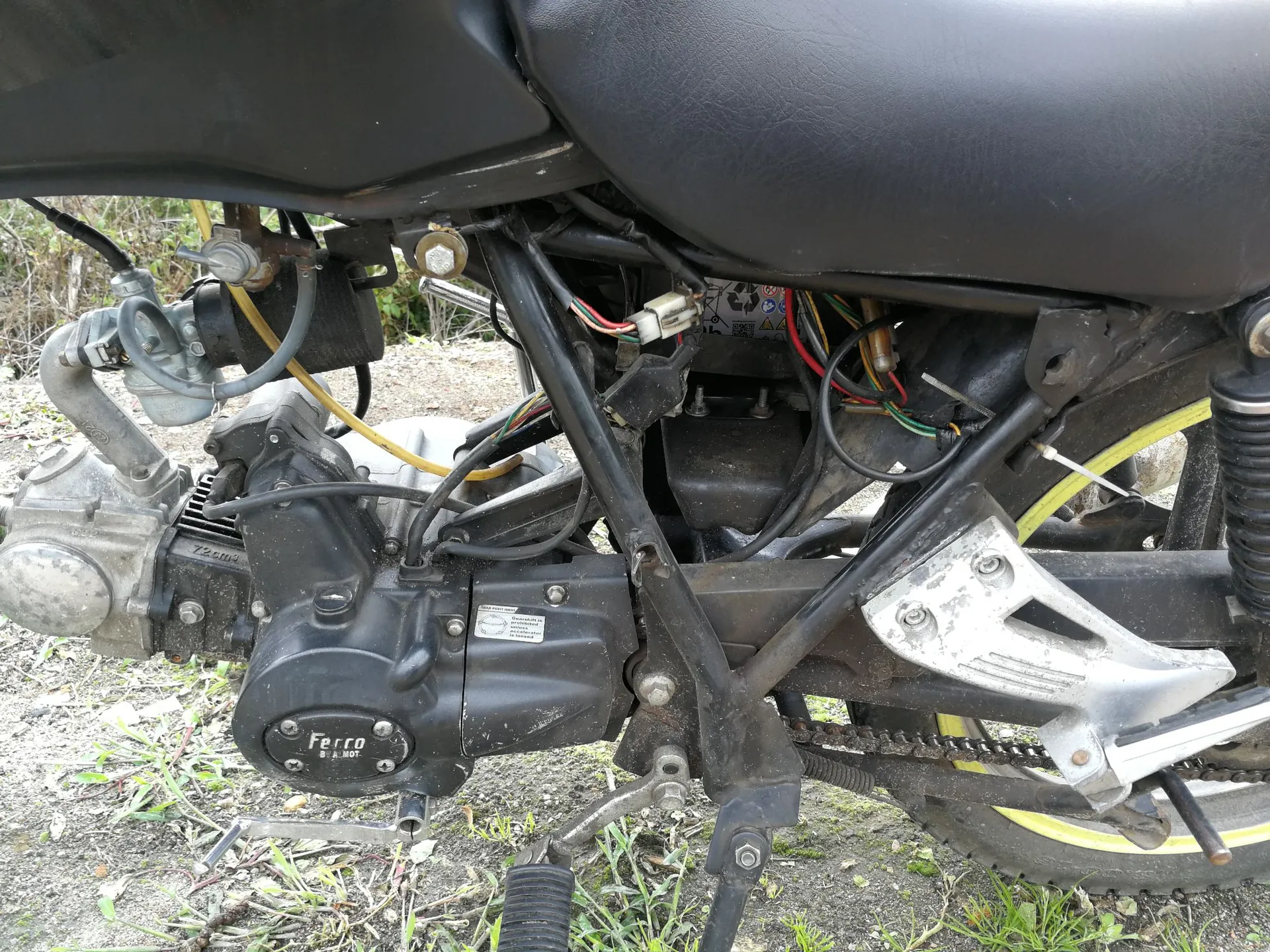 Zbliżenie na silnik i ramę motocykla marki Ferro. Widoczna jest czarna obudowa silnika z białym napisem 'Ferro BE A HOT', łańcuch napędowy, stopka oraz elementy instalacji elektrycznej z kolorowymi przewodami. Motocykl stoi na piaszczystym podłożu z kępkami trawy.