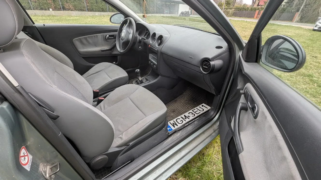 Wnętrze samochodu z otwartymi drzwiami od strony kierowcy. Widoczne są szare siedzenia, kierownica, deska rozdzielcza, dźwignia zmiany biegów oraz dywanik z tablicą rejestracyjną WGM-3EU1. Na drzwiach znajduje się naklejka zakazująca palenia. Poza samochodem trawa i ogrodzenie. W tle budynki i drzewa.