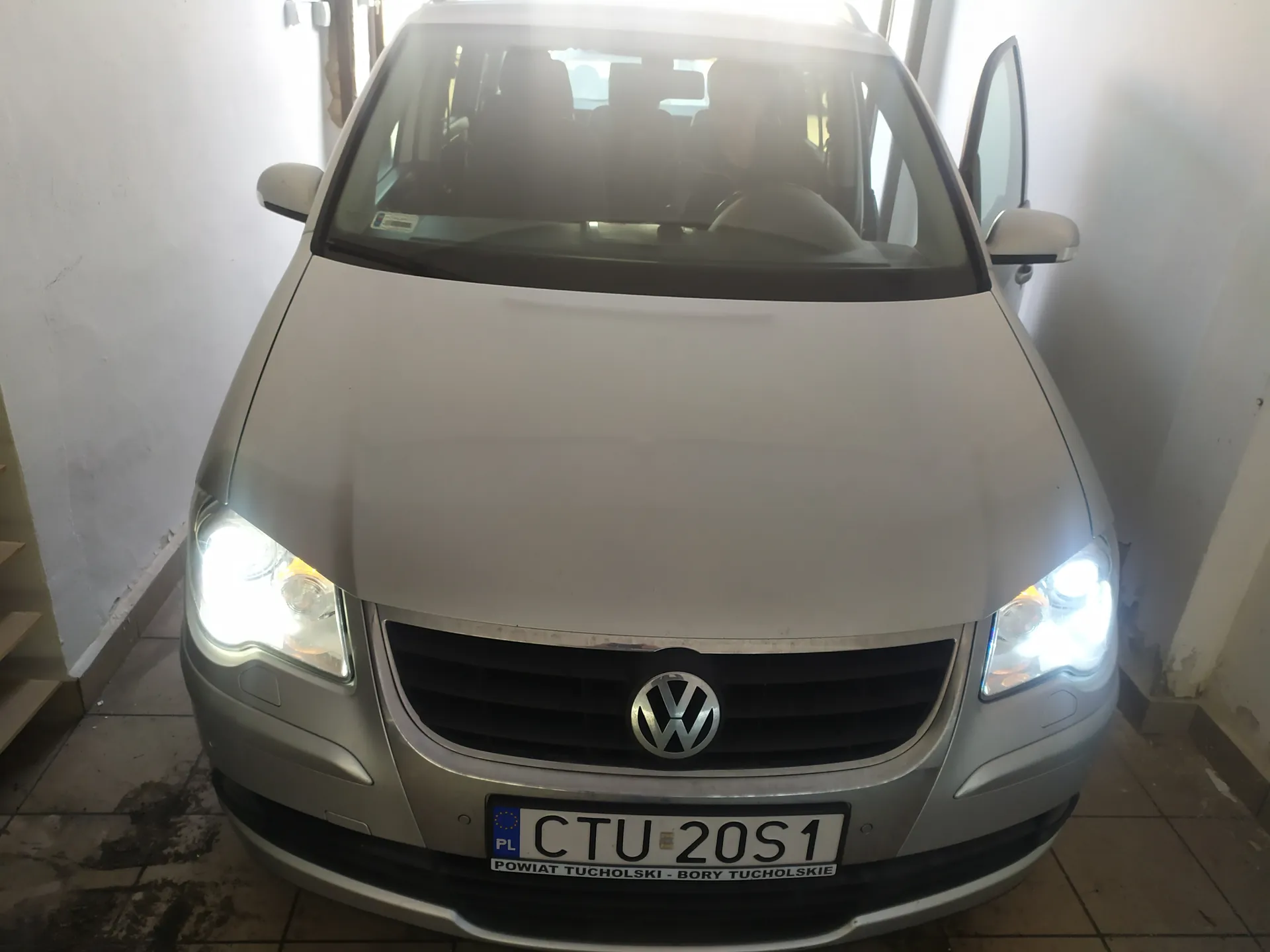 Srebrny samochód marki Volkswagen z przodu, z włączonymi reflektorami. Widoczna tablica rejestracyjna z napisem CTU 20S1 oraz podpisem POWIAT TUCHOLSKI - BORY TUCHOLSKIE. Drzwi po prawej stronie są otwarte. Samochód zaparkowany w pomieszczeniu z białymi ścianami i podłogą z kafelków.