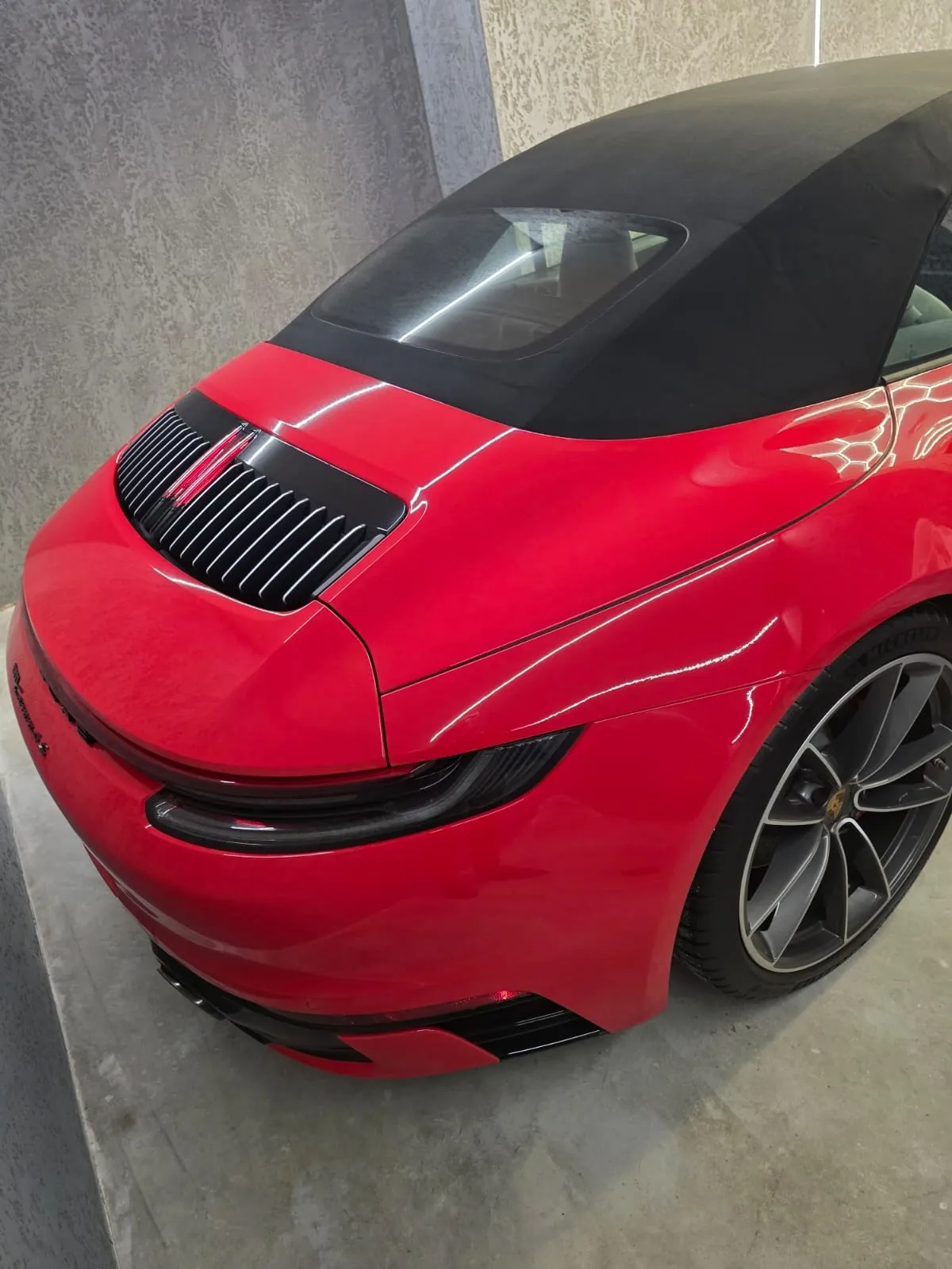 Czerwony samochód kabriolet z czarnym dachem, widok z przodu i boku. Na masce czarna atrapa chłodnicy z pionowymi listwami i czerwonym elementem w środku. Widoczne nowoczesne reflektory oraz felga z wieloramiennym wzorem. Samochód stoi w pomieszczeniu z betonowymi ścianami i podłogą.