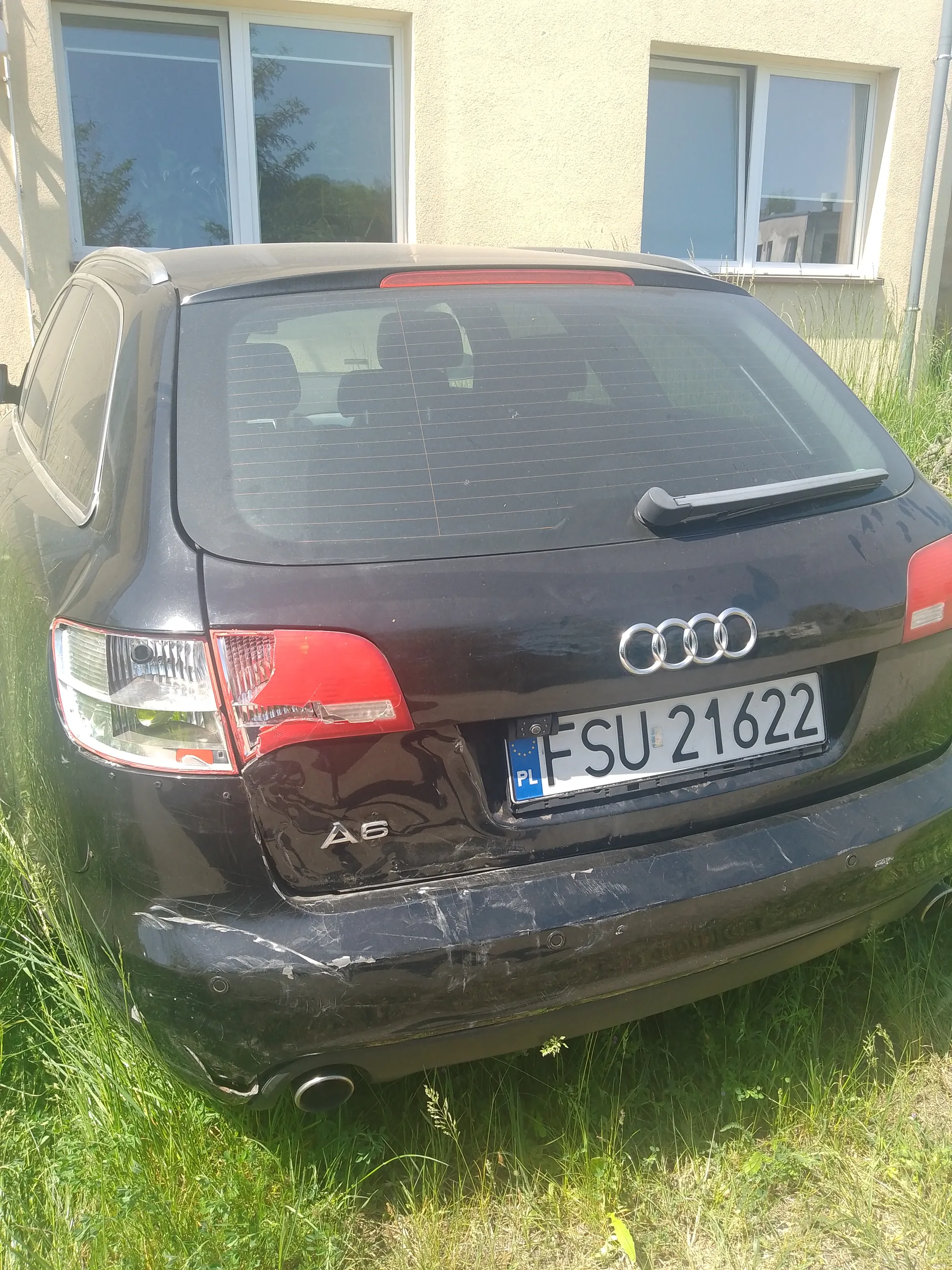 Tył czarnego samochodu Audi A6 z widocznym logo marki i oznaczeniem modelu A6. Tablica rejestracyjna z białym tłem, czarnym napisem FSU 21622 oraz niebieskim pasem z literami PL po lewej stronie. Samochód stoi na wysokiej trawie przed beżowym budynkiem z dwoma oknami. Widoczne są zadrapania na tylnym zderzaku i czerwone światła tylnego. Tylne szyby mają siatkę przewodów.
