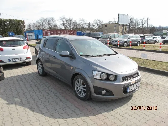Srebrny samochód Chevrolet Aveo zaparkowany na brukowanej powierzchni. W tle widoczne są inne pojazdy, w tym biały samochód z tablicą rejestracyjną KK 79481 oraz tablica KK 31425 na Chevroletu. Na pierwszym planie bruk, w tle budynki, drzewa bez liści, niebieskie kontenery i tablica reklamowa. W prawym dolnym rogu obrazu widoczna data 30/01/2010. Na parkingu stoją również czerwono-białe słupki.