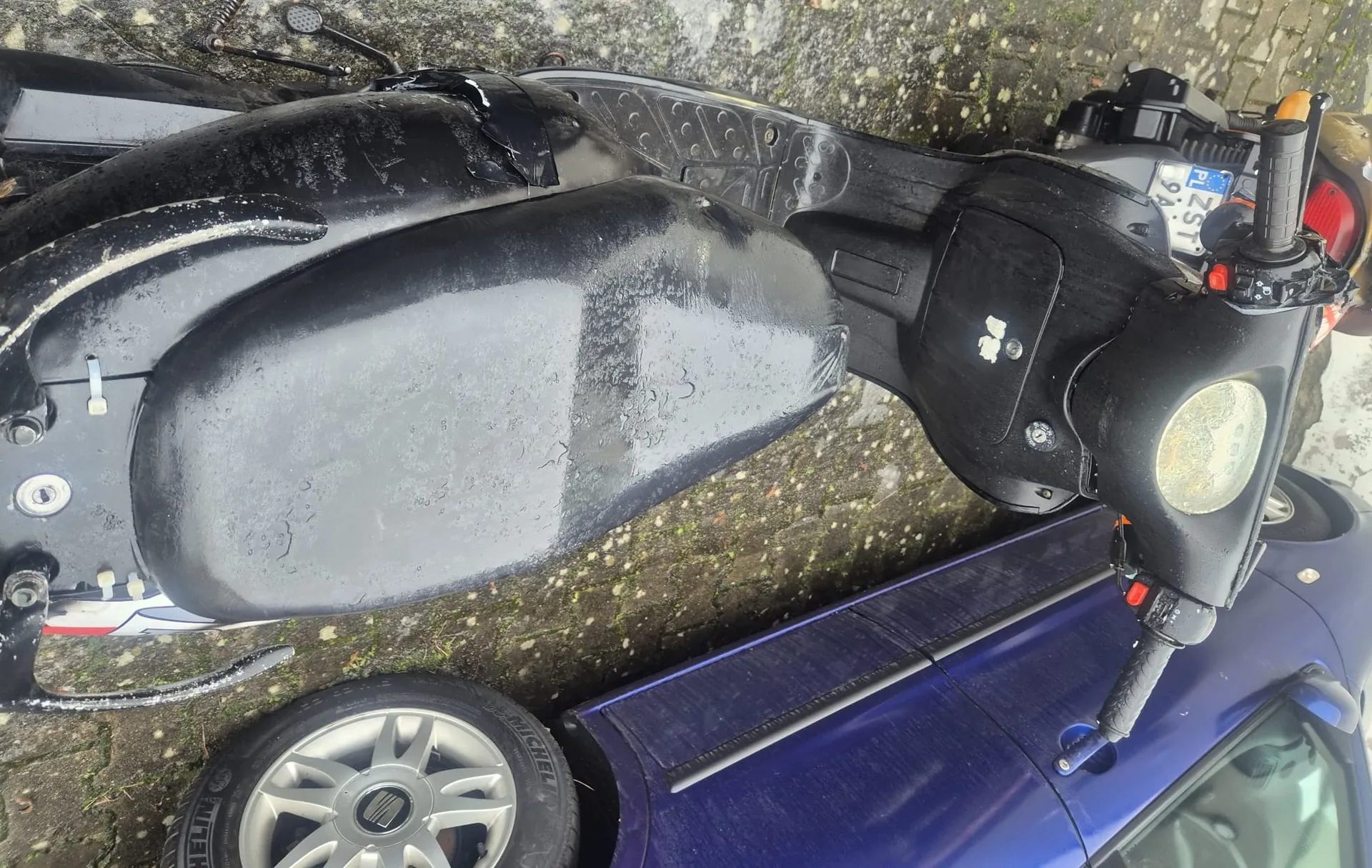 Czarny skuter z uszkodzonym siedzeniem owiniętym czarną taśmą, z widoczną tablicą rejestracyjną PL ZST 9A. Skuter ma reflektor, kierownicę z czarnymi uchwytami i czerwonymi przyciskami. Obok skutera stoi niebieski samochód z oponą Michelin. Tło stanowi brukowana powierzchnia z mchem i śladami wilgoci. Na skuterze widoczne są ślady zużycia i plamy.