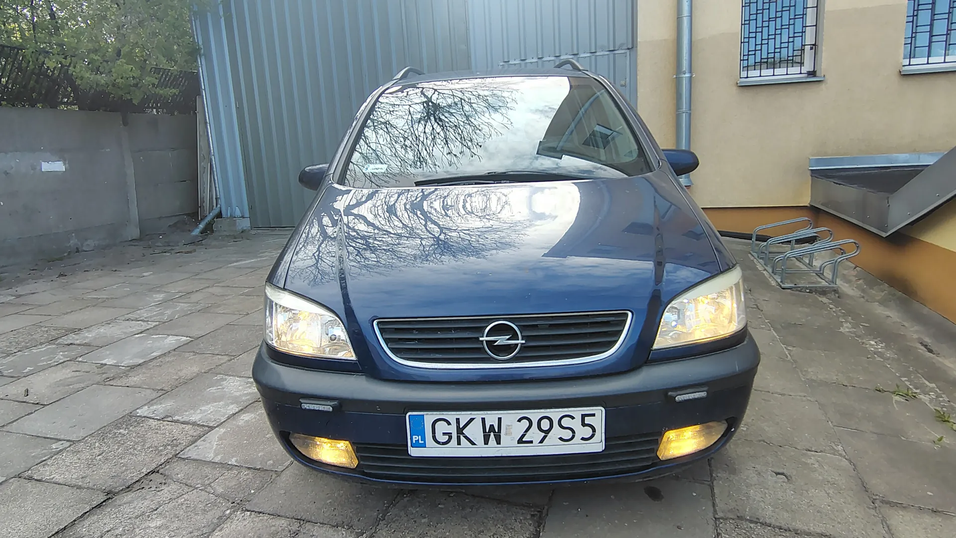 Niebieski samochód marki Opel z przodu, z widocznym logo na masce i tablicą rejestracyjną PL GKW 29S5. Samochód stoi na brukowanym podwórku przed budynkiem z metalową bramą i żółtymi ścianami. W tle widoczne są drzewa, betonowy mur oraz metalowe stojaki na rowery. Reflektory samochodu są włączone, a na masce odbijają się gałęzie drzew.