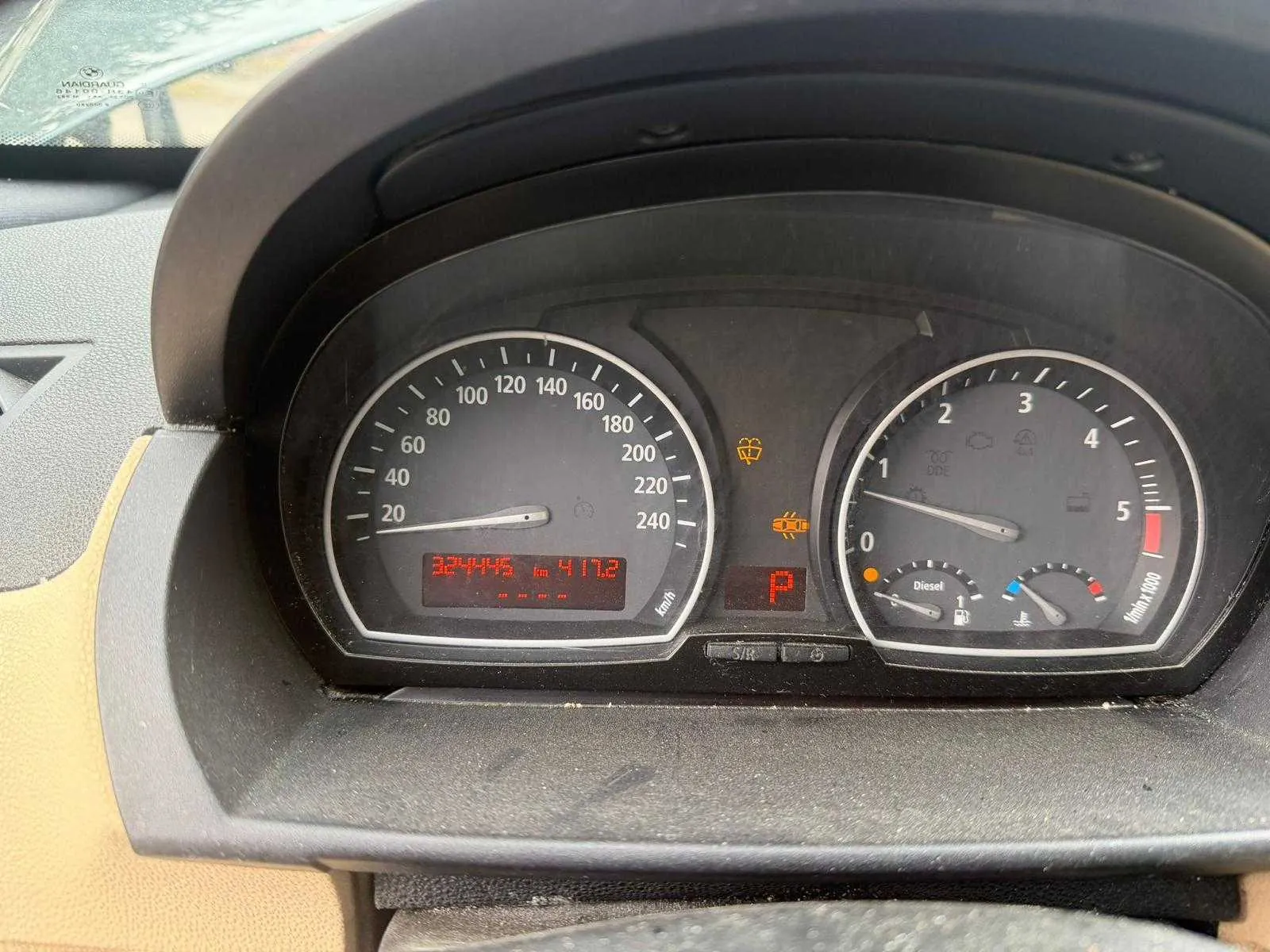 Zbliżenie na deski rozdzielcze samochodu. Po lewej stronie prędkościomierz z zakresem do 240 km/h, wskazówka na 0, cyfrowy wyświetlacz pokazujący 324445 km i 417,2. Po prawej tachometr z zakresem do 5 (x1000 obr./min), wskazówka na 0. W centralnej części czerwony symbol "P" (parking), pomarańczowe ikony ostrzegawcze (m.in. pas bezpieczeństwa, kontrola silnika). Widoczny napis "Diesel" oraz wskaźniki paliwa i temperatury. Część kierownicy i przedniej szyby widoczna na brzegach obrazu. Tekst i elementy są czytelne, ustawione pionowo.