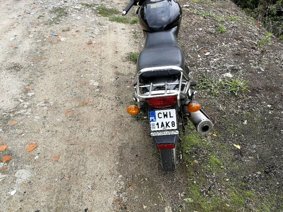 Widok z tyłu na czarny motocykl zaparkowany na piaszczystej drodze. Motocykl posiada srebrny bagażnik z tyłu oraz polską tablicę rejestracyjną o numerze CWL 1AK8.