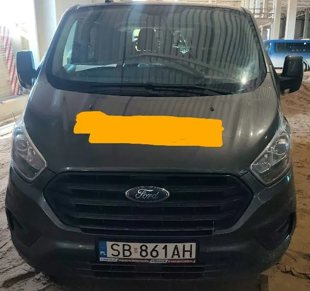 Widok z przodu ciemnoszarego vana marki Ford. Pojazd posiada czarny grill z logo Ford na środku oraz tablicę rejestracyjną o numerze SB 861AH z oznaczeniem PL. Część maski samochodu jest zakryta pomarańczowym polem. Auto znajduje się wewnątrz jasnego pomieszczenia, prawdopodobnie hali lub garażu.
