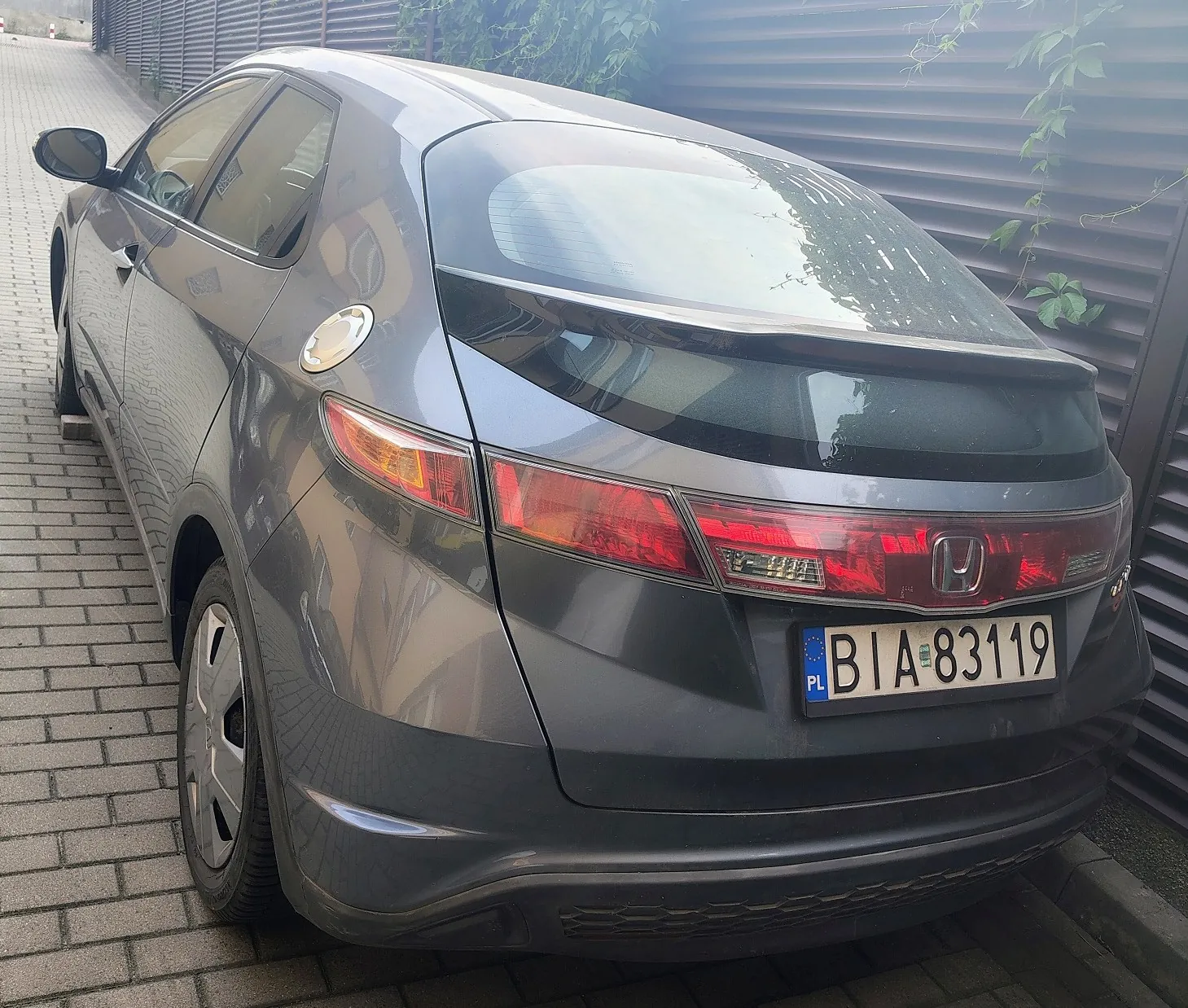 Tylny lewy profil szarego samochodu marki Honda Civic zaparkowanego na kostce brukowej obok ciemnego ogrodzenia. Widoczna jest tablica rejestracyjna o treści BIA 83119 z oznaczeniem PL.