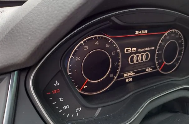 Zbliżenie na cyfrowy zestaw wskaźników w samochodzie Audi Q5 quattro. Po lewej stronie znajduje się duży okrągły prędkościomierz, po prawej mniejszy obrotomierz. Na środku widnieje napis 'Q5 quattro' oraz logo Audi składające się z czterech pierścieni. W górnej części ekranu widoczna jest data '29.4.2026', a na dole przebieg '84980 km' oraz czas '0:00'. W lewym dolnym rogu znajduje się wskaźnik temperatury z zaznaczoną wartością 130°C.