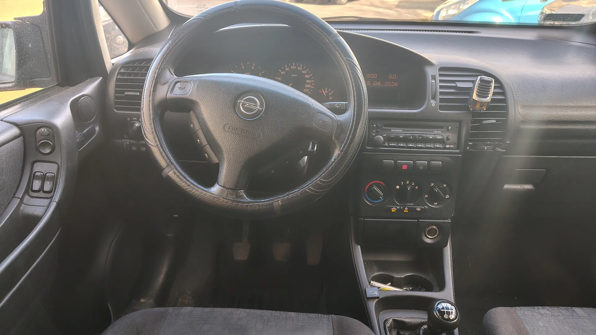 Wnętrze samochodu z widocznym kierownicą marki Opel, tablicą rozdzielczą z licznikami prędkości i obrotomierzem, wyświetlaczem cyfrowym pokazującym godzinę 8:00, wartość 6.0 oraz datę 18.04.2026. W centralnej konsoli znajduje się radio, przyciski klimatyzacji z oznaczeniami 1-2-3-4, dźwignia zmiany biegów z oznaczeniami 1-5 i R oraz przedmioty w uchwycie. Widoczne są również czarne siedzenia, pedały oraz fragment niebieskiego samochodu za przednią szybą.