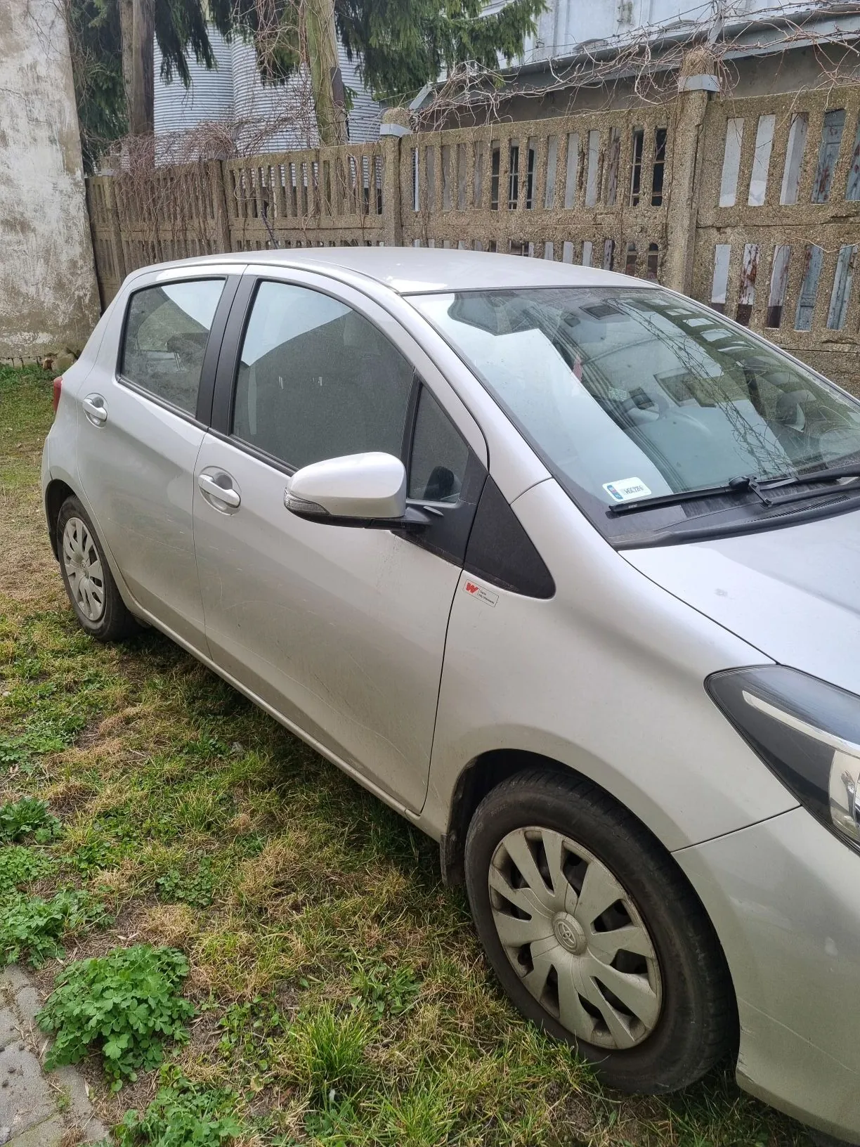 Srebrny samochód zaparkowany na trawie. Widoczne przednie koło, lusterko boczne, przednia szyba z naklejką. W tle drewniane ogrodzenie i fragment budynku. Trawa z zielonymi i suchymi plamami.