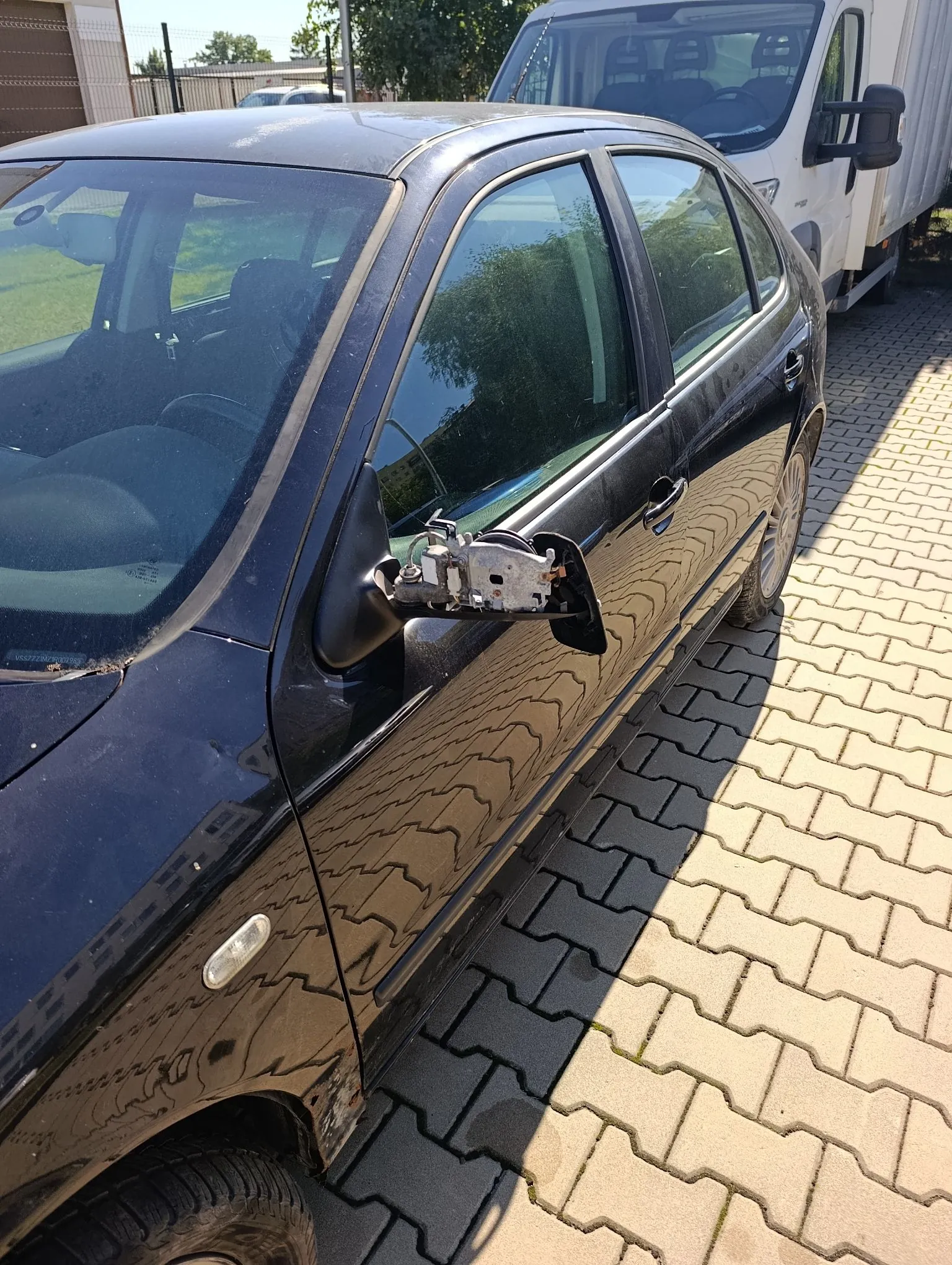 Czarny samochód osobowy zaparkowany na kostce brukowej. Widoczny jest uszkodzony prawy lusterko boczne, w którym brakuje obudowy i szkła, odsłaniając wewnętrzny mechanizm. W tle widoczny jest biały samochód dostawczy.