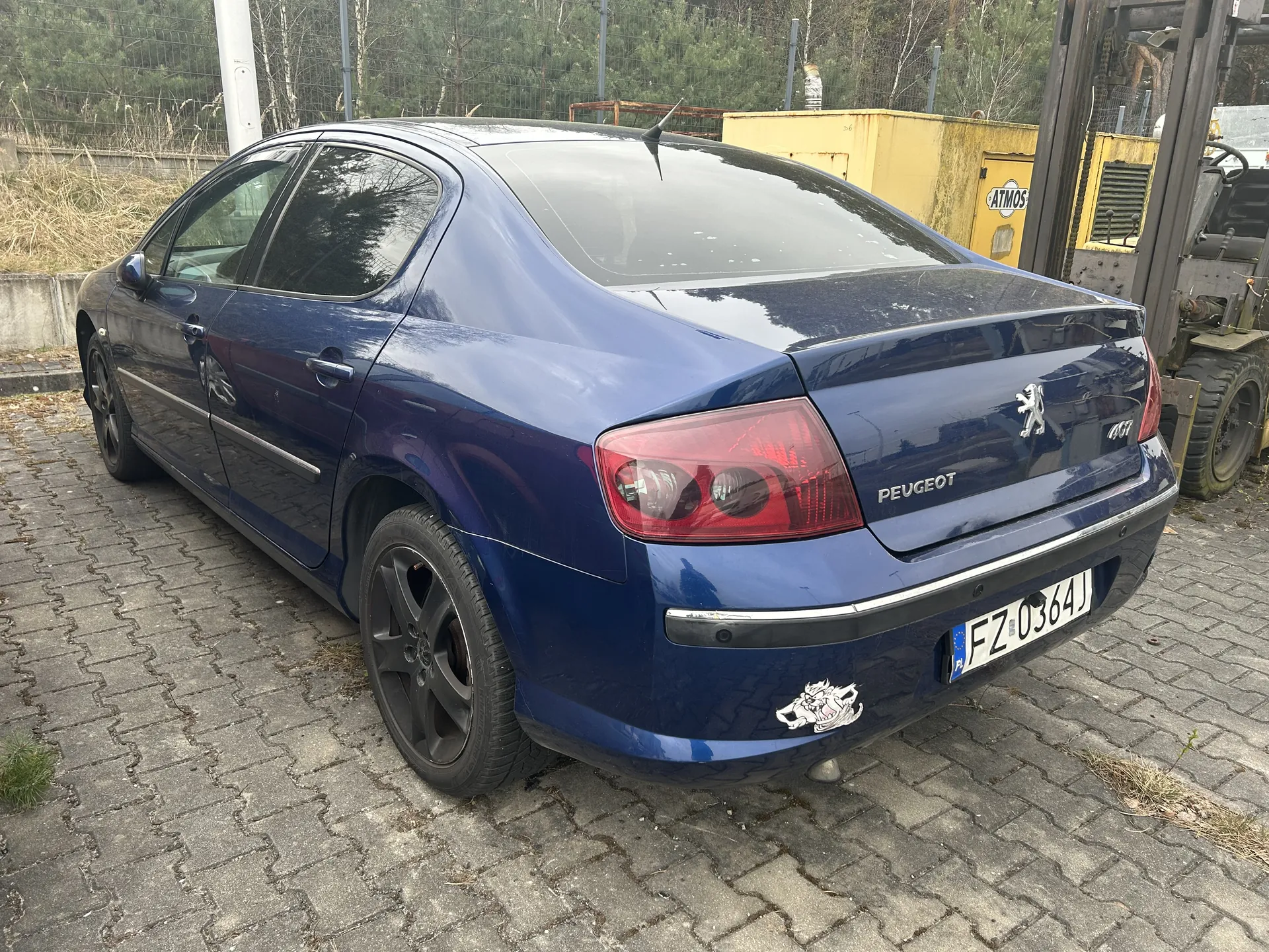 Niebieski samochód osobowy marki Peugeot 407 z widocznym tyłem, tablicą rejestracyjną FZ 0364J oraz naklejką na zderzaku. Po prawej stronie żółty kontener z napisem ATMOS i fragment maszyny przemysłowej. Tło stanowi ogrodzenie, suche trawy i drzewa. Samochód stoi na brukowanej powierzchni.
