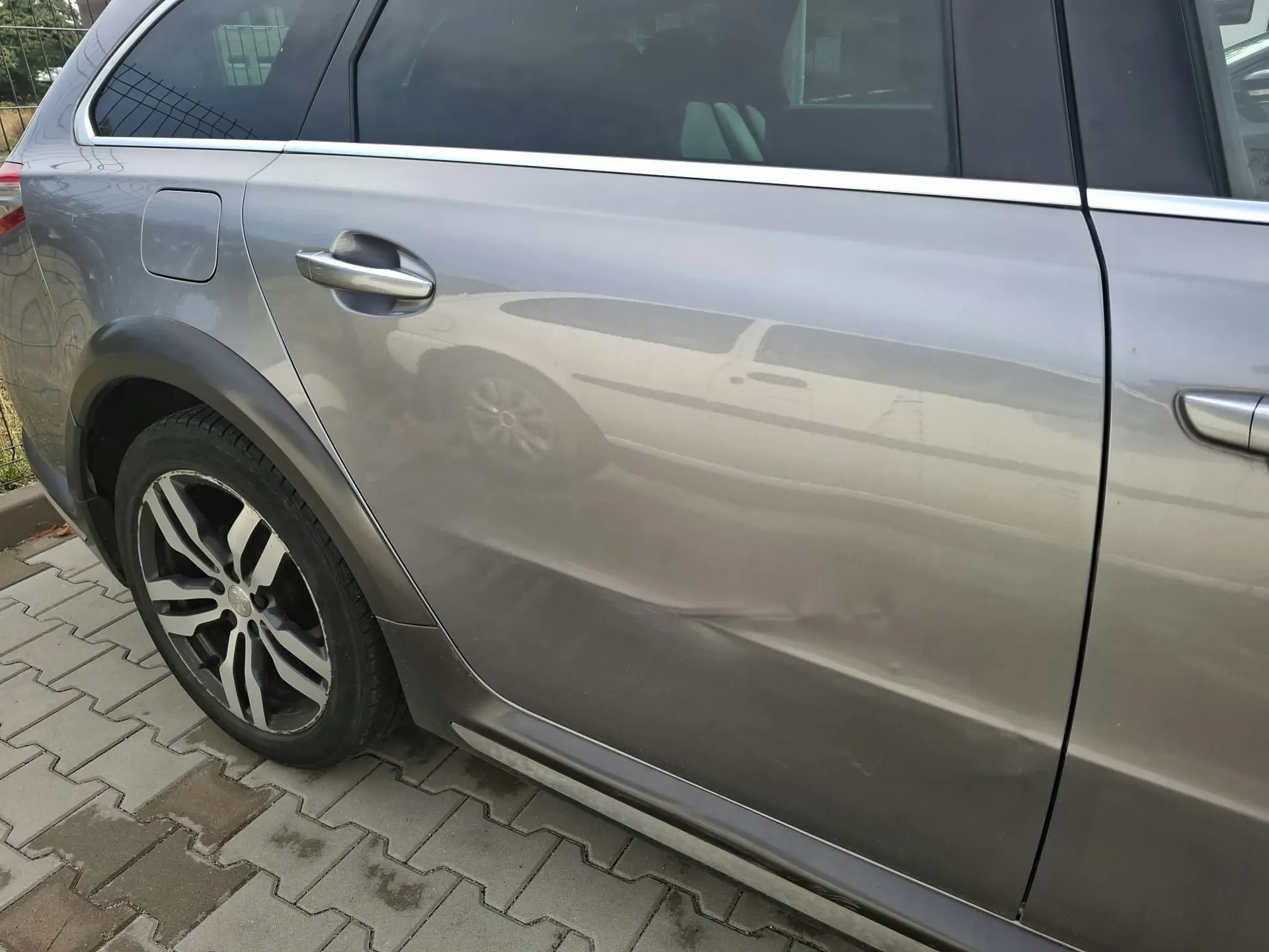 Zbliżenie na szary samochód typu kombi, ukazujące wgniecenie na tylnych drzwiach od strony pasażera. Widoczna jest felga aluminiowa z oponą oraz fragment nadwozia z klamką. Samochód zaparkowany jest na kostce brukowej.
