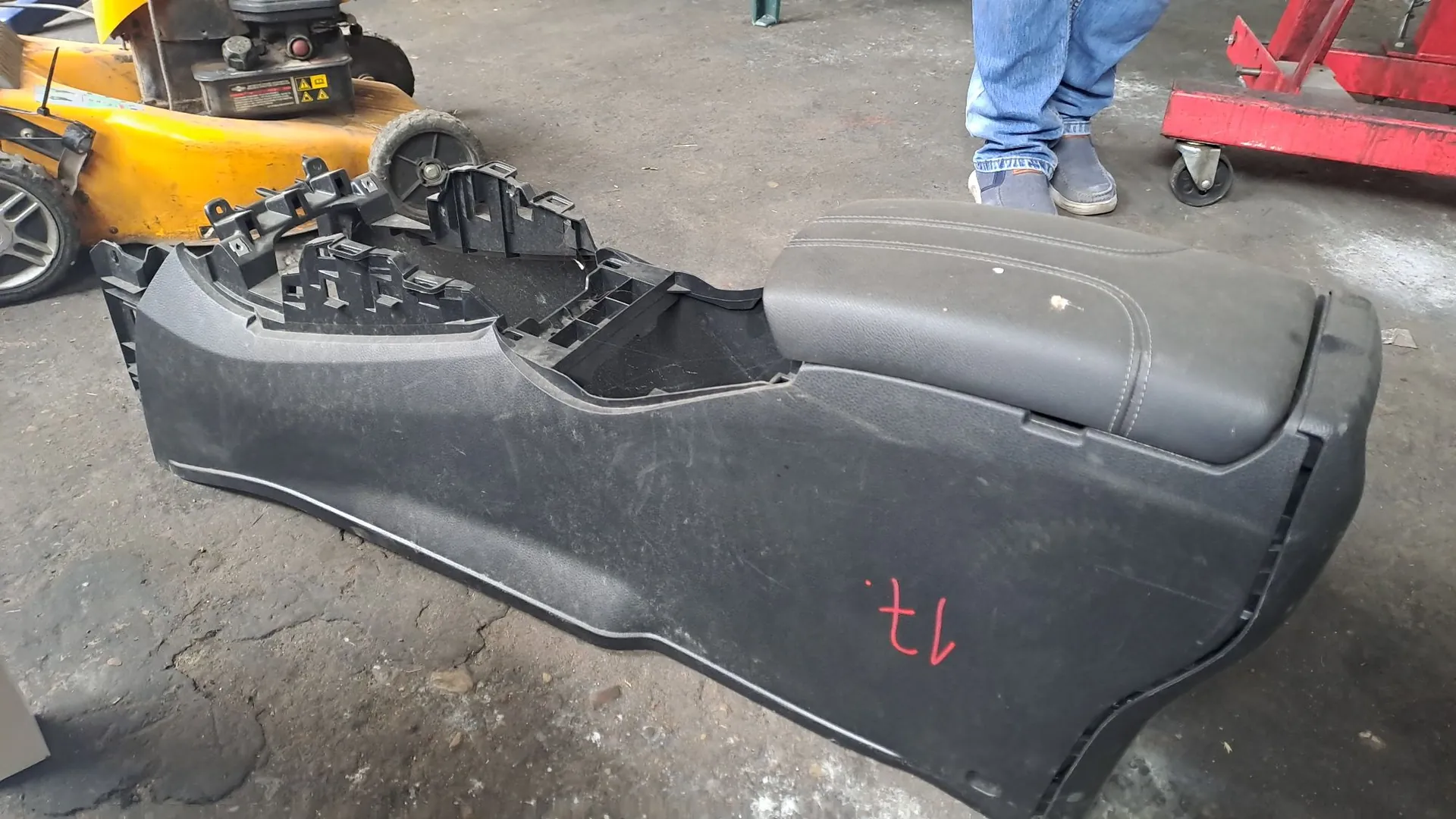 Czarna, plastikowa konsola środkowa z samochodu, leżąca na betonowej podłodze warsztatu. Element posiada podłokietnik z ekoskóry oraz wgłębienia na przyciski i uchwyty. Na boku konsoli widnieje czerwony napis '17. tl'. W tle widoczny jest żółty wózek warsztatowy, nogi osoby w niebieskich spodniach oraz fragment czerwonego podnośnika samochodowego.