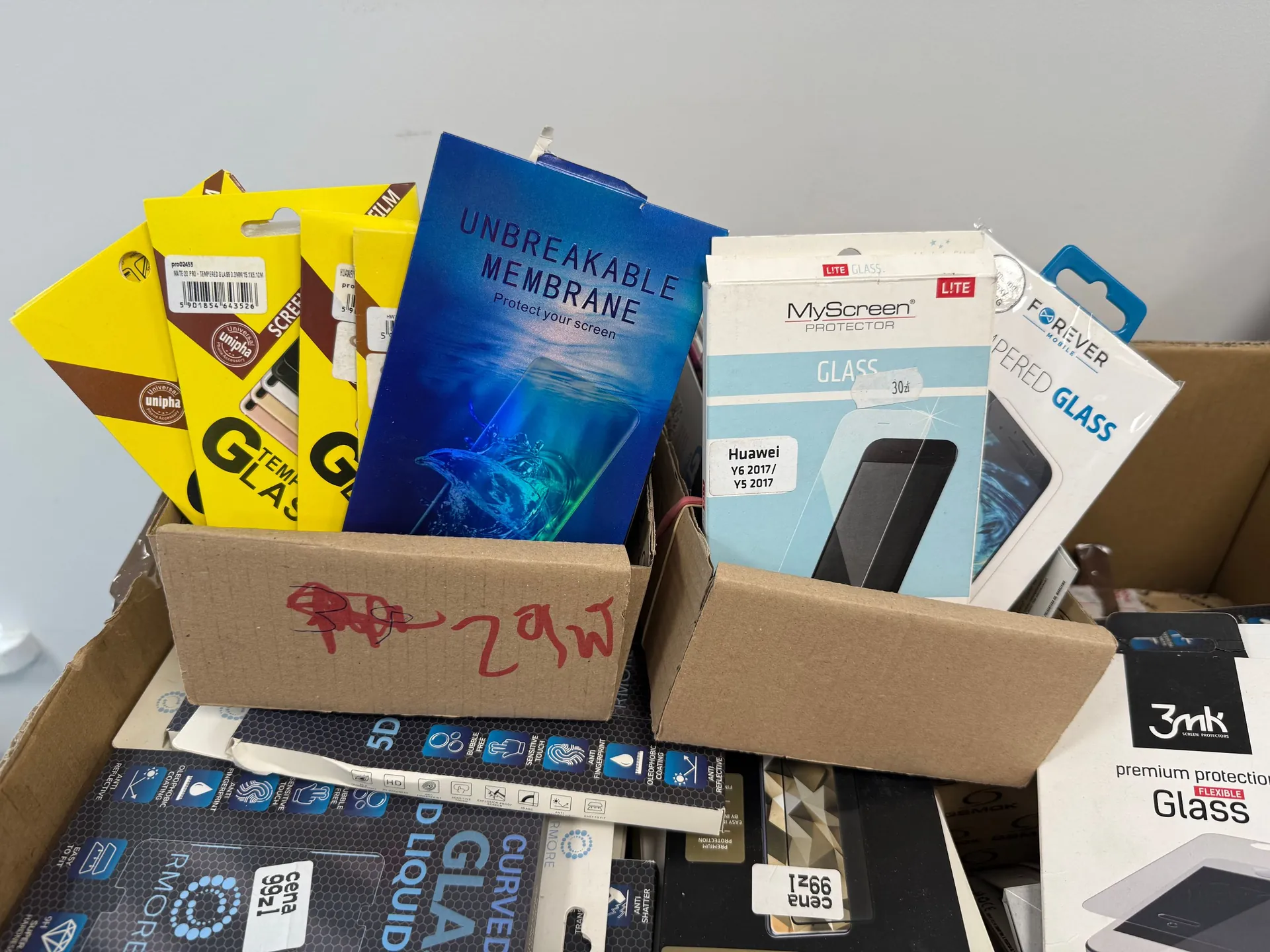 Kartonowe pudełko wypełnione różnymi opakowaniami szkieł ochronnych do telefonów. Widoczne są kolorowe pudełka z napisami: 'UNBREAKABLE MEMBRANE Protect your screen', 'MyScreen PROTECTOR GLASS', 'FOREVER GLASS' oraz '3mk premium protection Glass'. Po lewej stronie znajduje się żółte opakowanie z napisem 'GLASS'. Wewnątrz pudełka widoczne są również inne elementy opakowań z tekstem 'CURVED GLASS' oraz 'LIQUID'.
