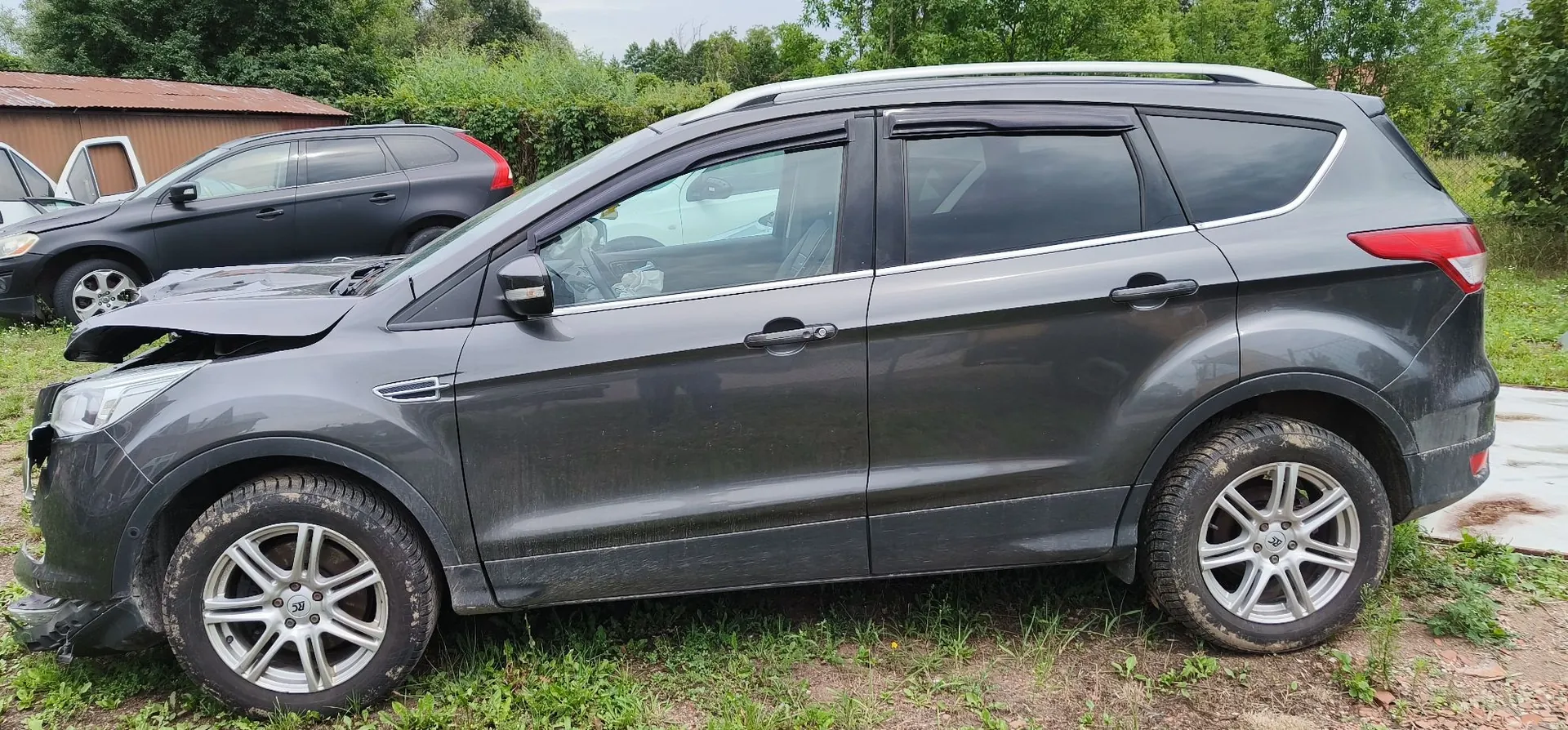 Profil ciemnoszarego samochodu typu SUV z widocznymi uszkodzeniami przodu, w tym wgniecioną maską i zniszczonym zderzakiem. Pojazd stoi na trawiastym podłożu. W tle widoczny jest inny, czarny samochód oraz brązowy budynek i zielone drzewa.