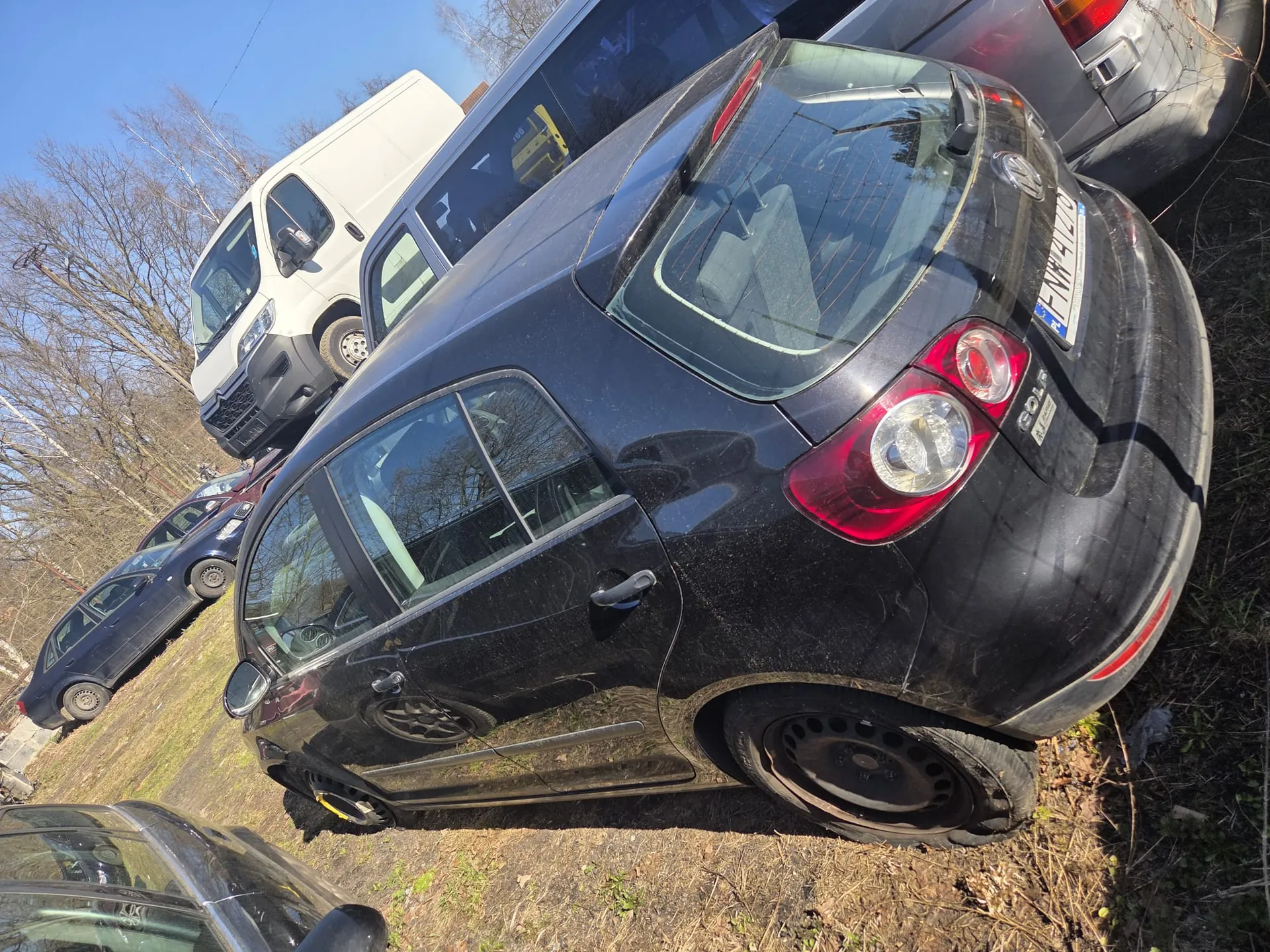 Kilka samochodów zaparkowanych na trawiastym obszarze z suchymi plamami. W centrum czarny Volkswagen Golf z widoczną tablicą rejestracyjną FNH 2179. W tle biały furgon, ciemny sedan oraz drzewa bez liści pod jasnym niebem.