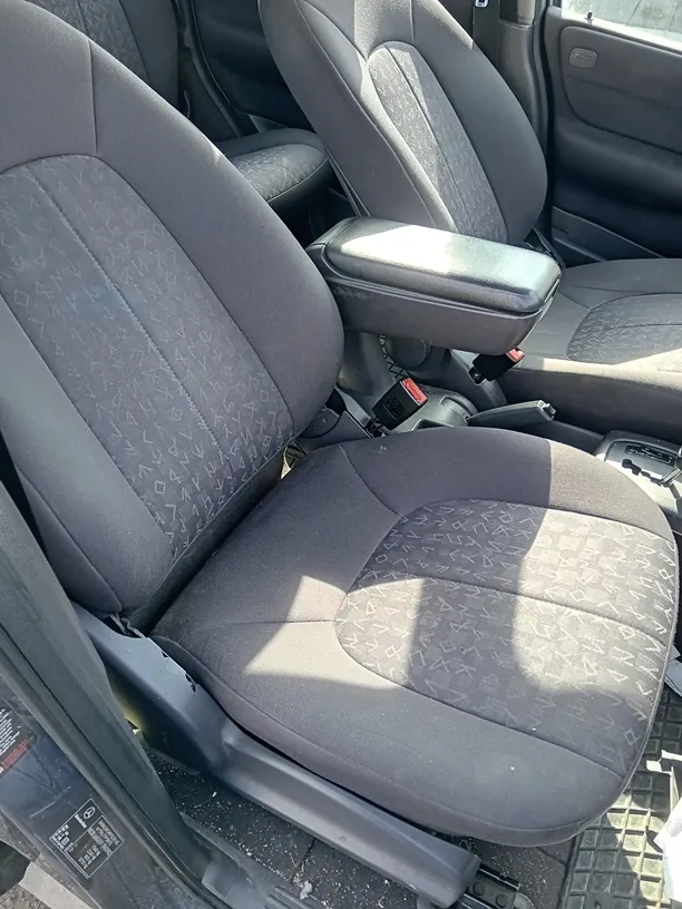 Wnętrze samochodu z widocznymi przodowymi siedzeniami w kolorze ciemnoszarym z geometrycznym wzorem. Siedzenia mają podłokietnik między nimi z otwartym schowkiem. Drzwi po lewej stronie są otwarte, na ich krawędzi widoczna jest czarna etykieta z białym tekstem. Podłoga pokryta jest gumową wykładziną. Światło słoneczne rzuca cienie na siedzenia.