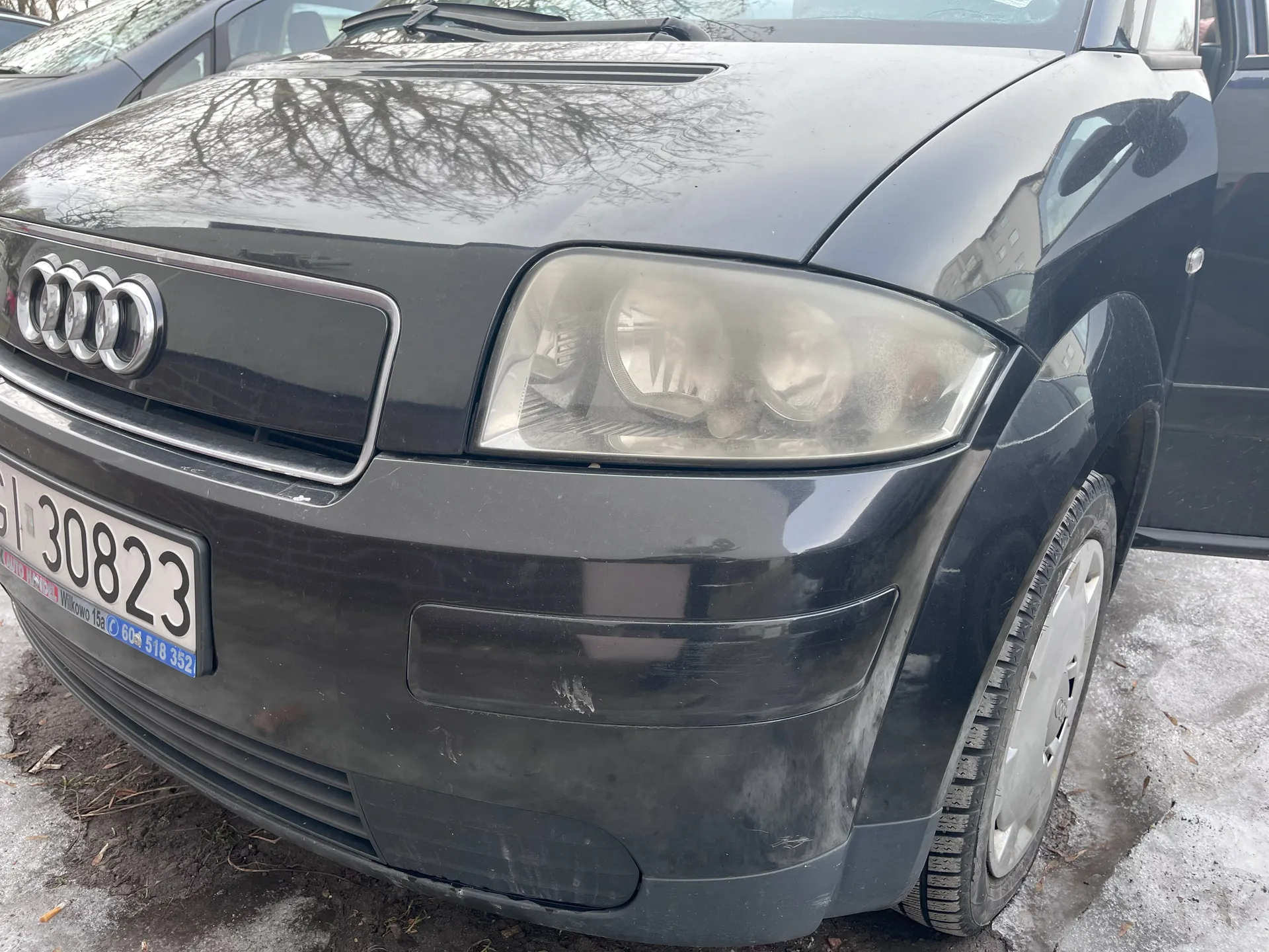 Czarny samochód marki Audi, widok z przodu. Widoczna maska z czterema pierścieniami w logo, reflektor, tablica rejestracyjna z numerem 30823 oraz napisami "Włocławek 15a" i "604 518 352". Drzwi pasażera są otwarte. Tło: śnieżna powierzchnia i fragment innego pojazdu. Na masce odbijają się gałęzie drzew.