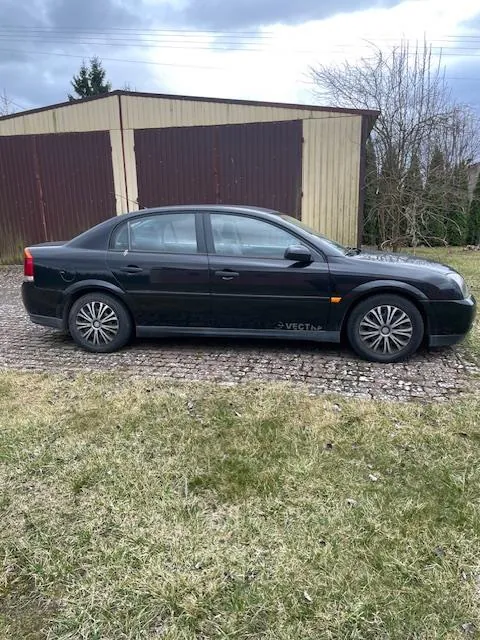 Czarny samochód osobowy typu sedan marki Opel Vectra zaparkowany na żwirowym podjeździe. Pojazd znajduje się przed budynkiem garażowym o beżowo-brązowej elewacji. W tle widoczne są suche drzewa, zielona trawa na pierwszym planie oraz zachmurzone niebo. Na boku samochodu widnieje napis "VECTRA".
