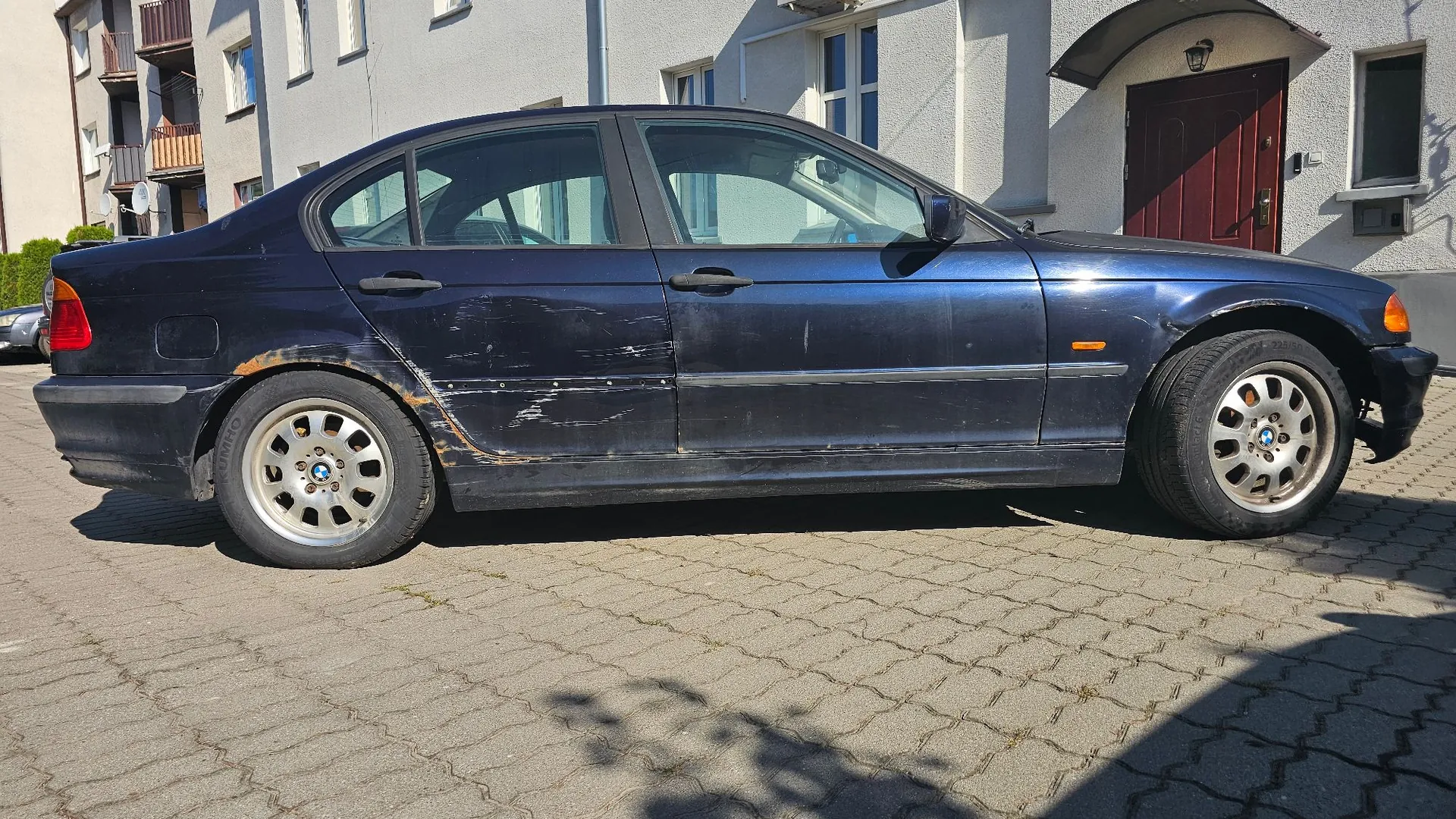 Profil ciemnoniebieskiego samochodu marki BMW zaparkowanego na kostce brukowej przed jasnym budynkiem. Na bocznych drzwiach i nad tylnym kołem widoczne są liczne zarysowania oraz ogniska korozji.