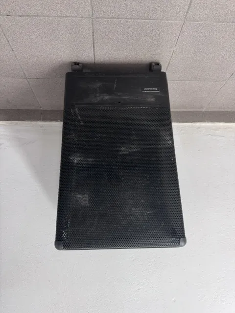 Czarny głośnik kolumnowy z metalową siatką ochronną, stojący w narożniku pomieszczenia przy białej ścianie i szarej płytkowej podłodze. Na obudowie widoczny jest napis 'Harman'.