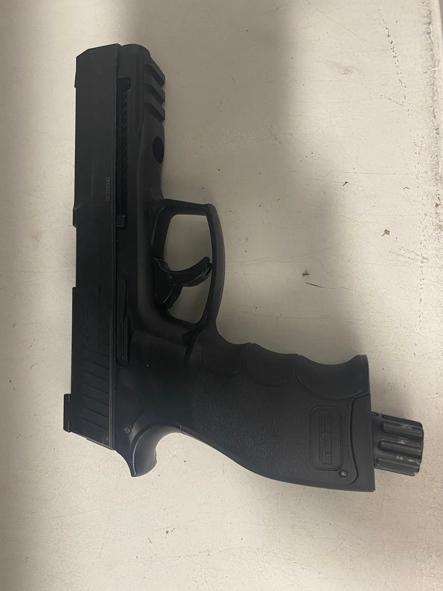 Czarny pistolet leżący na jasnej powierzchni z widocznymi zadrapaniami i drobnymi plamami. Na rękojeści widoczny napis 'T4E', a na ramie pistoletu numer seryjny 'DC225343'. Pistolet jest ułożony poziomo z lufą skierowaną w lewo.