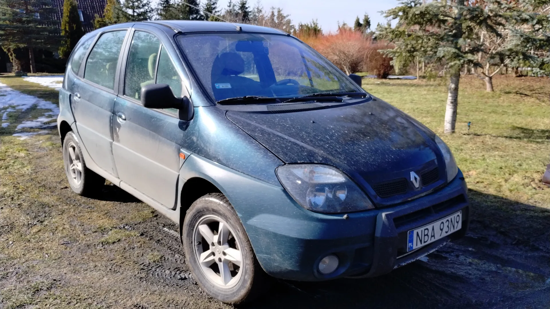 Niebieski samochód osobowy marki Renault, typu hatchback, zaparkowany na trawiastym i błotnistym terenie. Widoczna tablica rejestracyjna NBA 93N9 z oznaczeniem PL. Na tle drzewa, krzewy, dom z ciemnym dachem oraz fragmenty śniegu po lewej stronie. Samochód pokryty jest kurzem, a na przedniej szybie widoczny jest biały naklejka. Tło stanowi jasne niebo i zielona trawa.