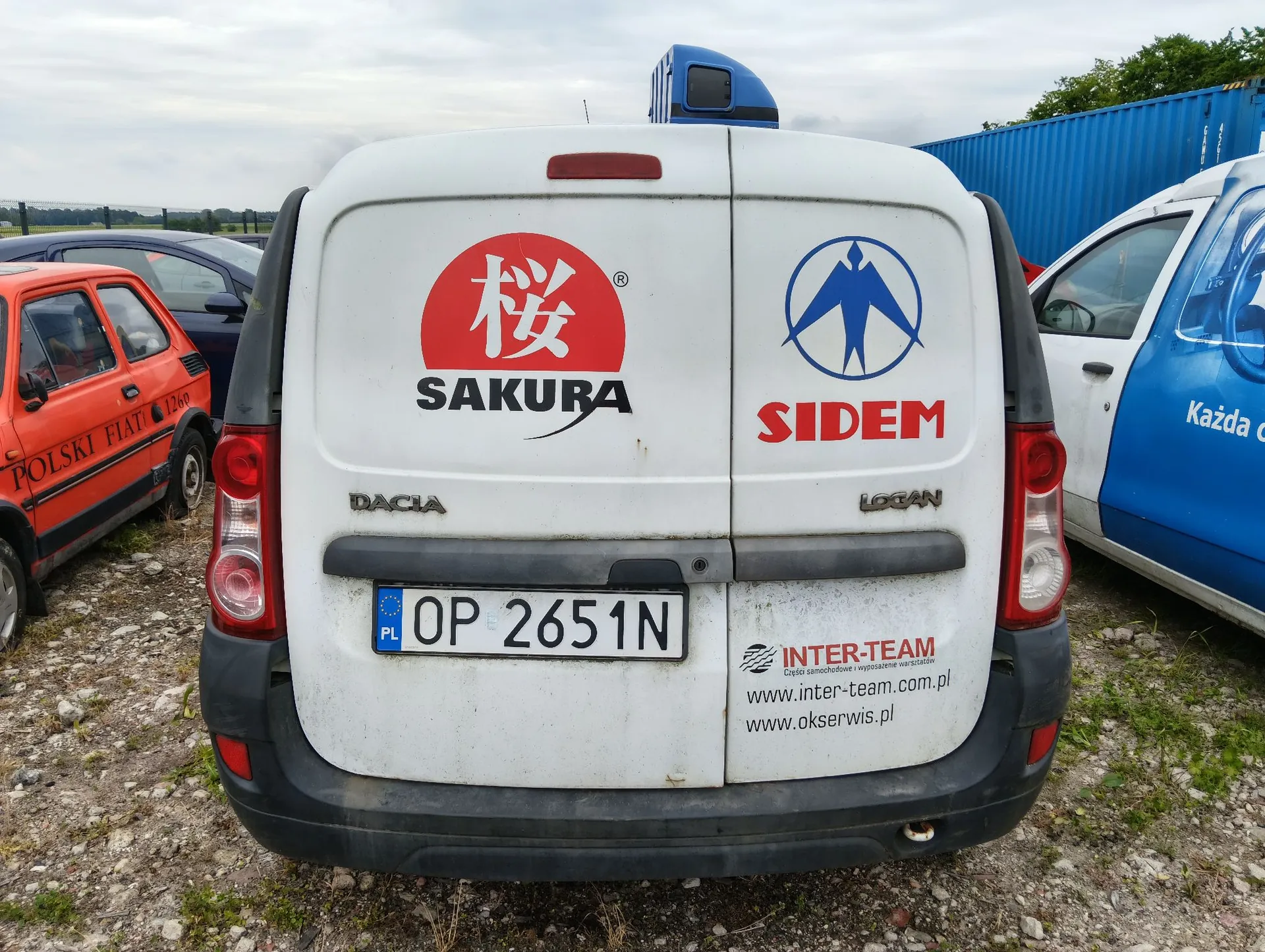 Tylny widok białego samochodu dostawczego marki Dacia Logan. Na tylnych drzwiach znajdują się dwa duże logo: po lewej czerwone koło z białym znakiem i napisem 'SAKURA', po prawej niebieski symbol ptaka w okręgu z czerwonym napisem 'SIDEM'. Poniżej znajduje się tablica rejestracyjna o numerze 'OP 2651N'. W prawym dolnym rogu widnieje naklejka z napisem 'INTER-TEAM Części samochodowe i wyposażenie warsztatów' oraz adresami stron internetowych 'www.inter-team.com.pl' i 'www.okserwis.pl'. Po lewej stronie stoi czerwony samochód z napisem 'POLSKI FIAT', a po prawej biało-niebieski samochód.