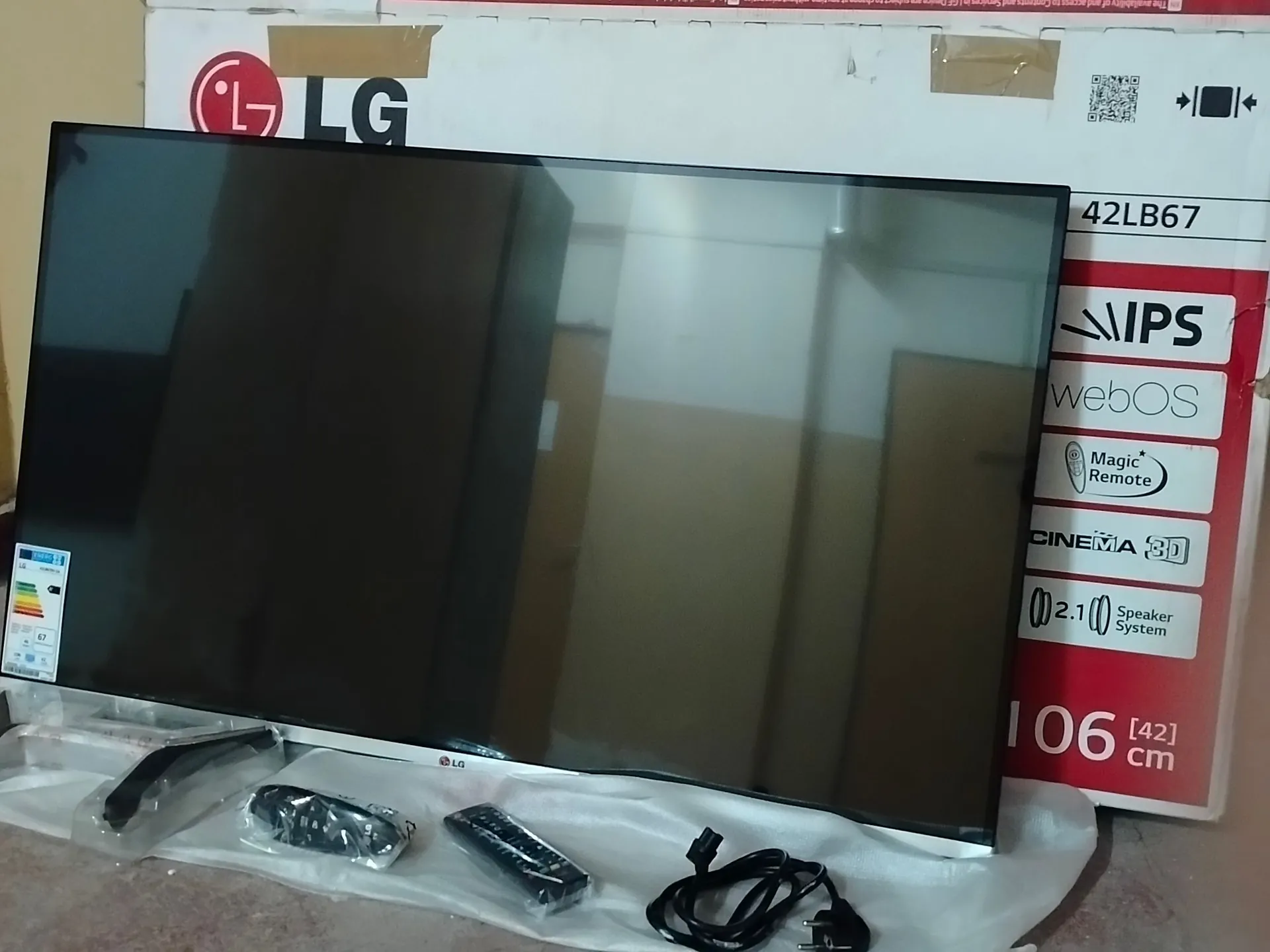 Czarny telewizor LG z serii 42LB67, ustawiony przed swoim oryginalnym białym opakowaniem. Na pudełku widoczne są logotypy LG, IPS, webOS, Magic Remote, Cinema 3D oraz napis '106 cm [42]'. Przed telewizorem na białej folii ochronnej leżą akcesoria: dwie nóżki do podstawy, pilot oraz kabel zasilający.