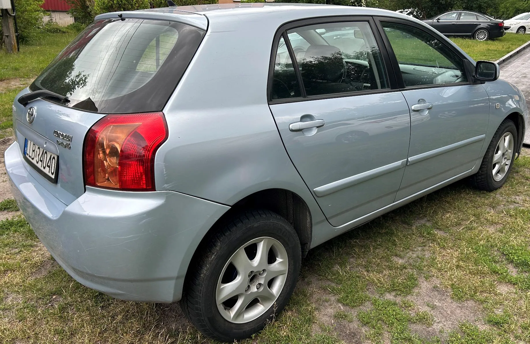 Jasnoniebieski samochód osobowy marki Toyota Corolla w wersji hatchback, zaparkowany na trawiastym podłożu. Zdjęcie przedstawia tył i prawy bok pojazdu. Samochód ma srebrne felgi aluminiowe i tablicę rejestracyjną o numerze LLB 34040.