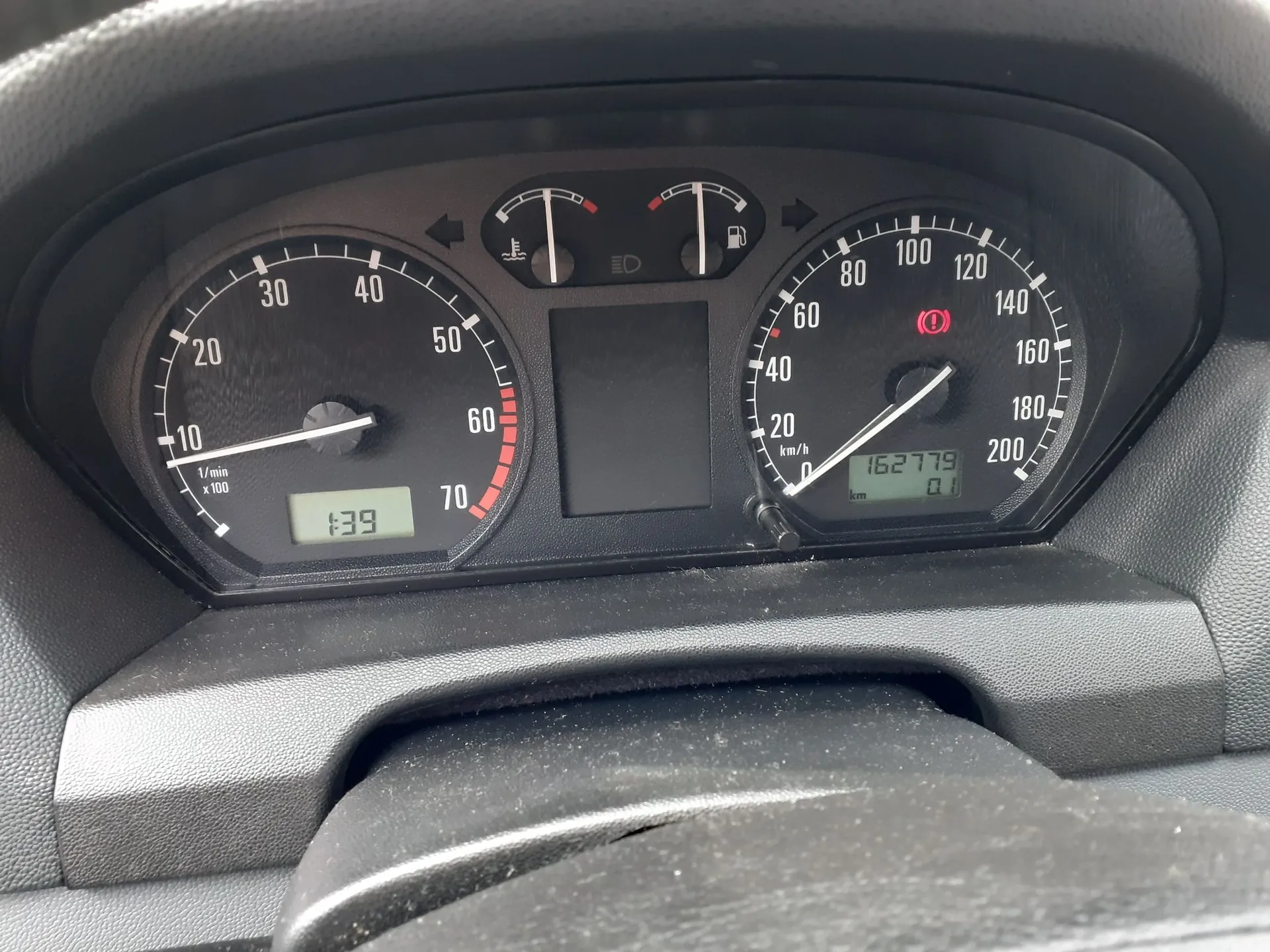 Zbliżenie na deskę rozdzielczą samochodu. Widoczny jest prędkościomierz po prawej stronie z maksymalną wartością 200 km/h i cyfrowym licznikiem przebiegu wskazującym 16277.9 km. Po lewej stronie znajduje się obrotomierz z podziałką od 10 do 70 (x100 1/min) oraz małym cyfrowym wyświetlaczem z liczbą 1:39. W górnej części środkowej znajdują się wskaźniki temperatury silnika i poziomu paliwa oraz kontrolka świateł. Na prędkościomierzu świeci się czerwona kontrolka układu hamulcowego.