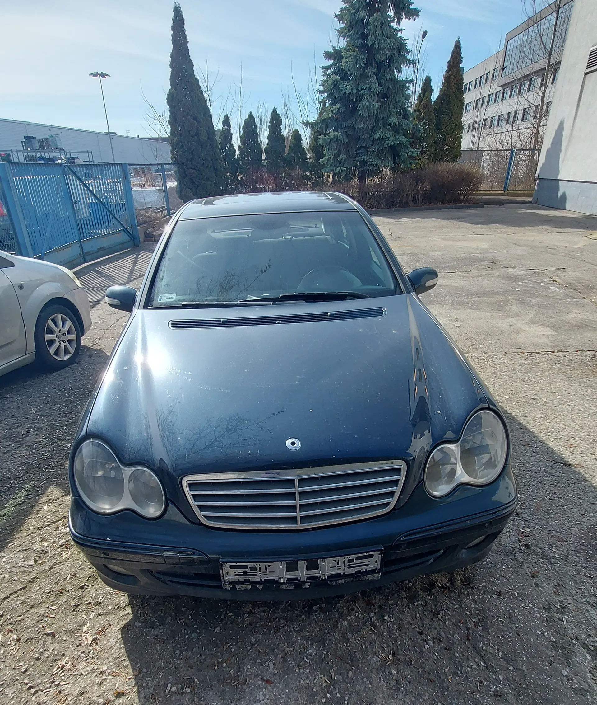 Ciemnoniebieski samochód marki Mercedes-Benz zaparkowany na podwórku z żwirem i betonem. Widoczna przód pojazdu z uszkodzoną tablicą rejestracyjną, reflektorami i charakterystyczną atrapą chłodnicy. Po lewej stronie częściowo widoczny srebrny samochód. W tle niebieska metalowa brama, wysokie drzewa iglaste, budynek z oknami oraz jasne niebo z nielicznymi chmurami.