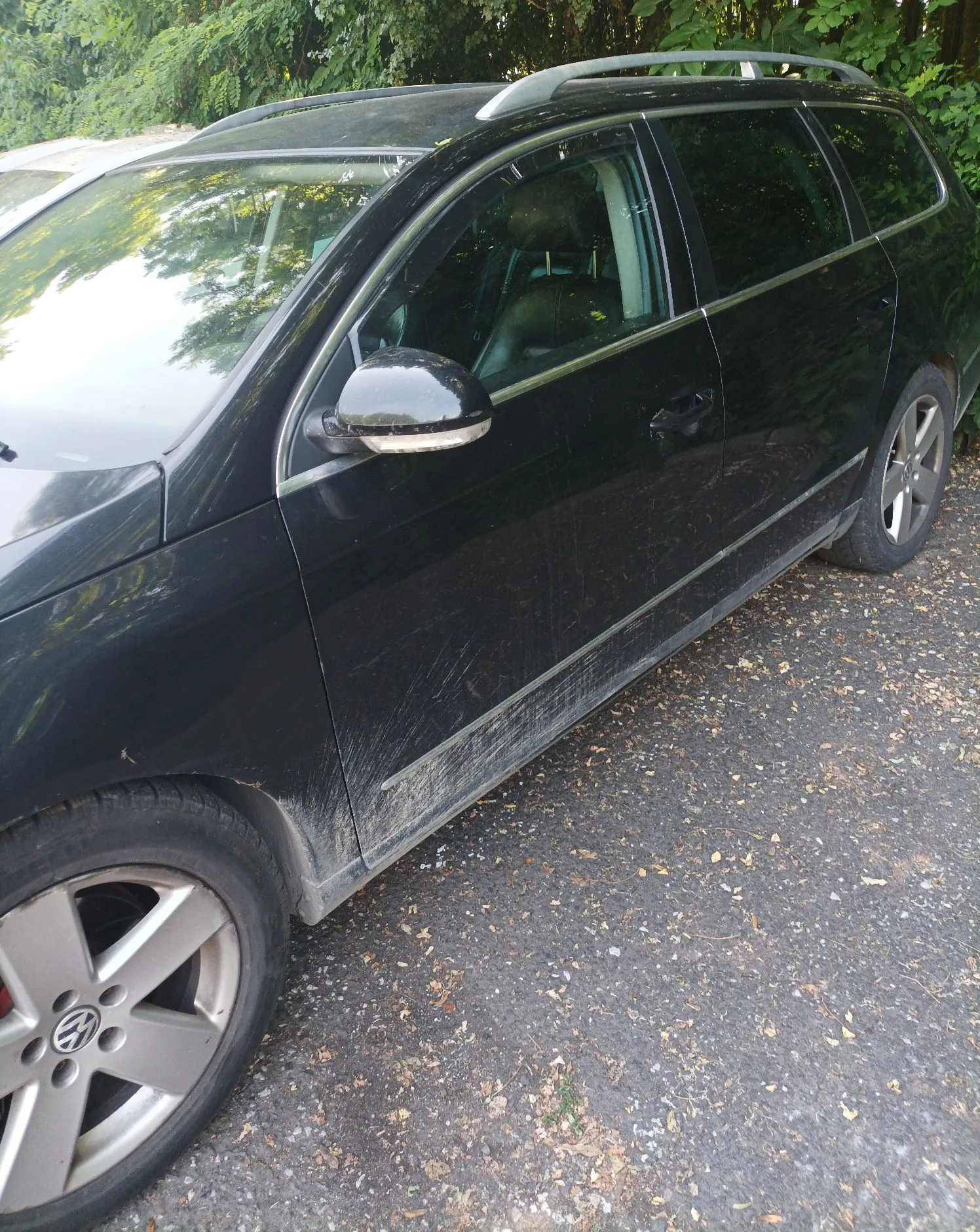 Czarny samochód typu kombi zaparkowany na żwirowym podłożu. Widoczny jest bok pojazdu z jednej strony, w tym przednie i tylne koło z felgami aluminiowymi z logo Volkswagena. Na dolnej części drzwi widoczne są zarysowania i zabrudzenia. W tle znajdują się zielone drzewa i krzewy.