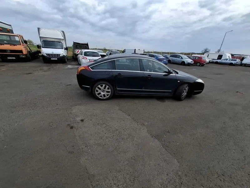 Czarny samochód osobowy typu sedan na pierwszym planie, zaparkowany na asfaltowym placu. W tle widoczne są różne pojazdy, w tym ciężarówki i inne samochody osobowe. Niebo jest zachmurzone.