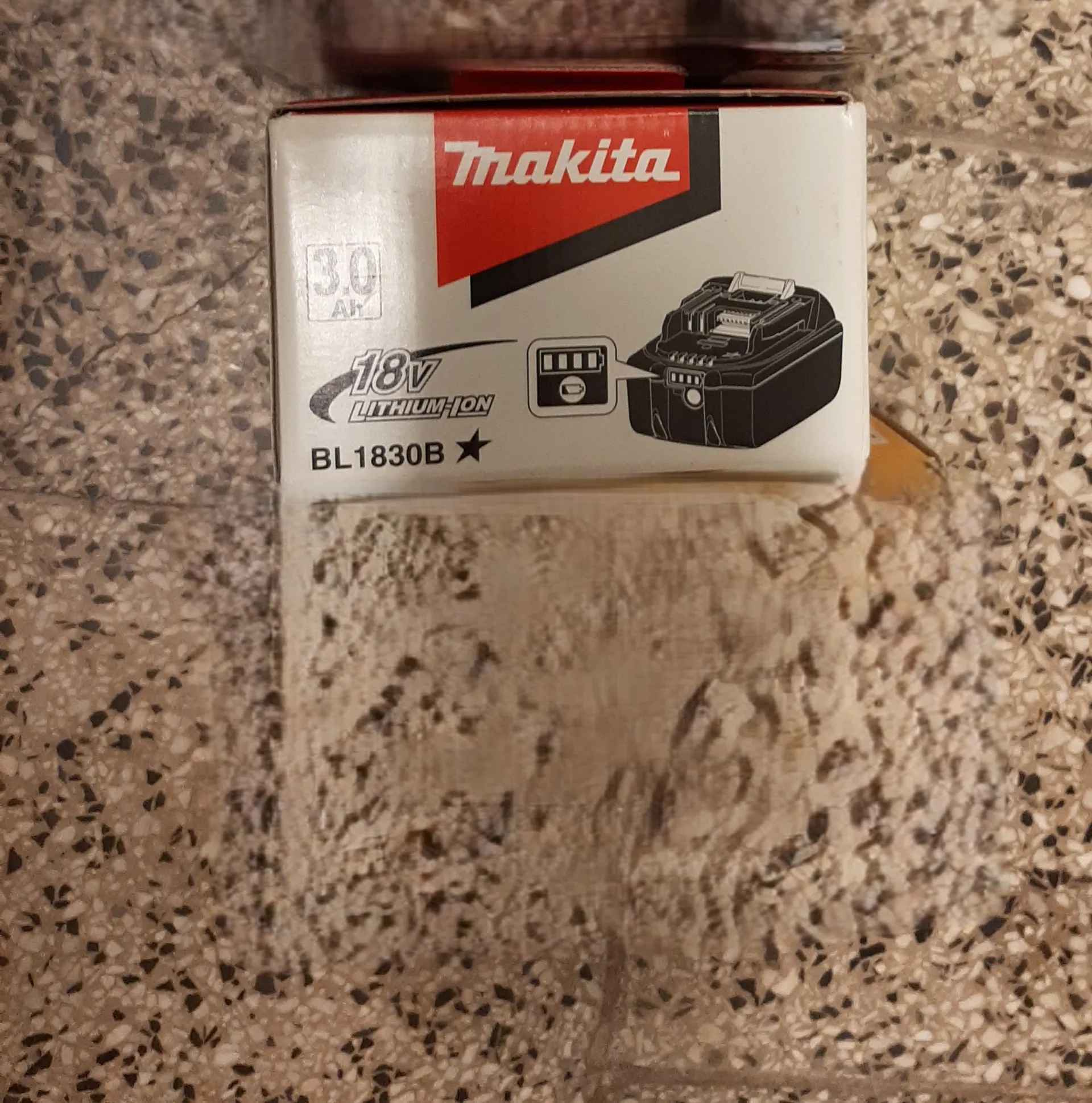 Białe pudełko z czerwonym logo Makita, na którym widnieją napisy 3.0 Ah, 18V LITHIUM-ION, BL1830B oraz czarno-biała grafika akumulatora. Pudełko leży na powierzchni z kropkowanym wzorem w odcieniach brązu i czerni.