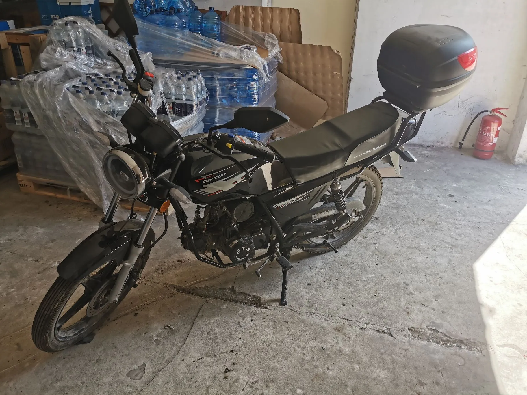 Czarny motocykl marki Barton zaparkowany w pomieszczeniu przypominającym magazyn. Motocykl posiada zamontowany czarny kufer z tyłu oraz okrągły reflektor z przodu. W tle widoczne są stosy plastikowych butelek z wodą owinięte folią stretch, kartonowe pudła oraz czerwona gaśnica na ścianie. Podłoże stanowi betonowa posadzka.